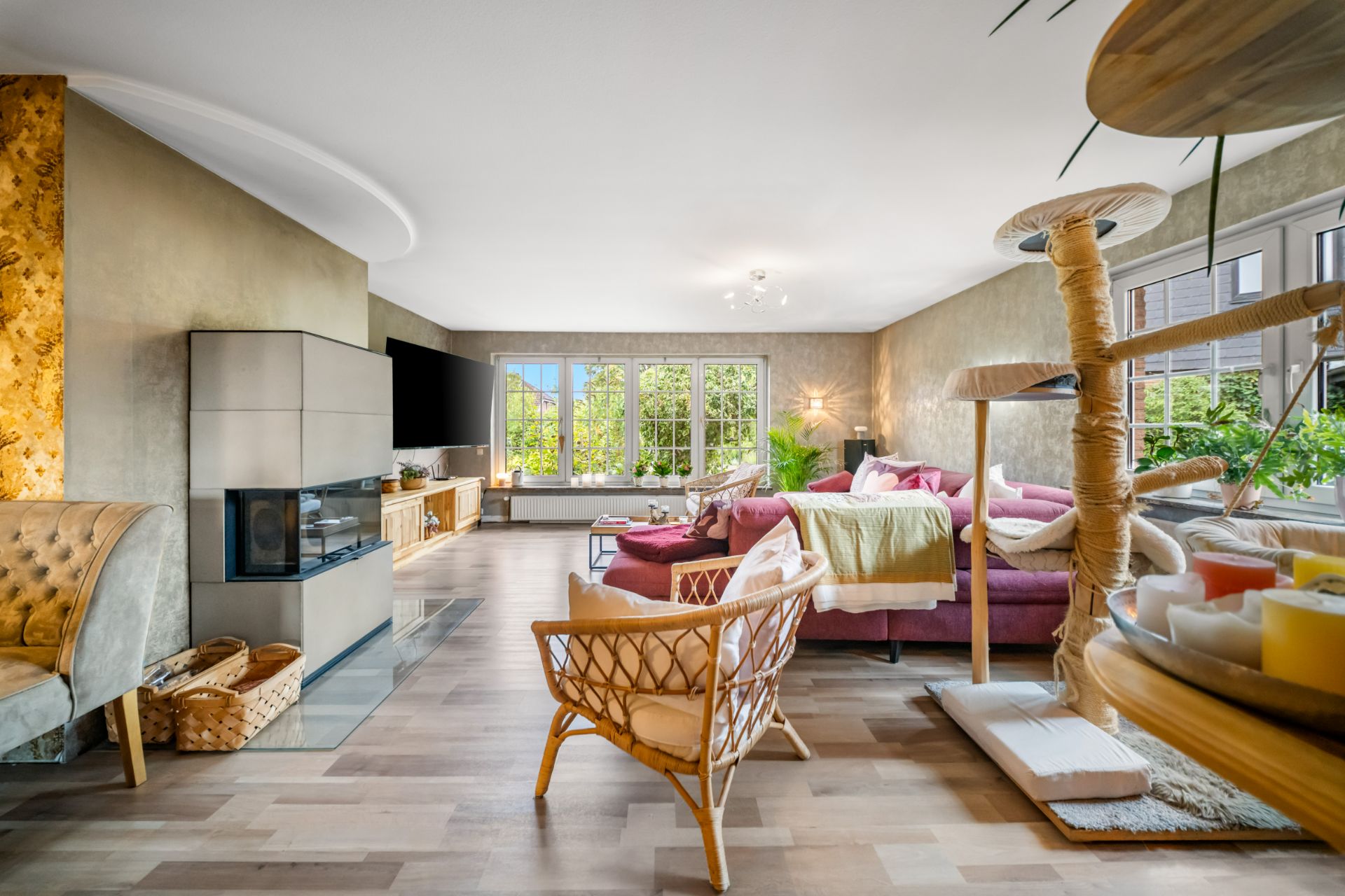 Helles Wohnzimmer mit Kamin, Sofa, Sessel, Fernseher und großen Fenstern mit Blick ins Grüne.