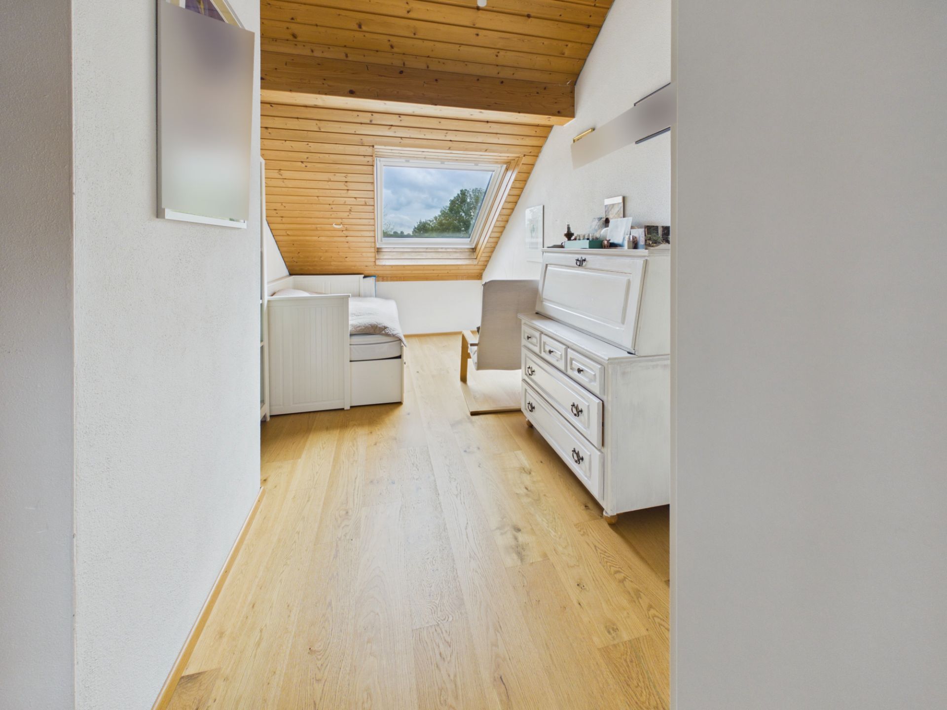 Helles Zimmer mit Holzboden, Dachschräge und großem Fenster, links ein Bett, rechts eine weiße Kommode.