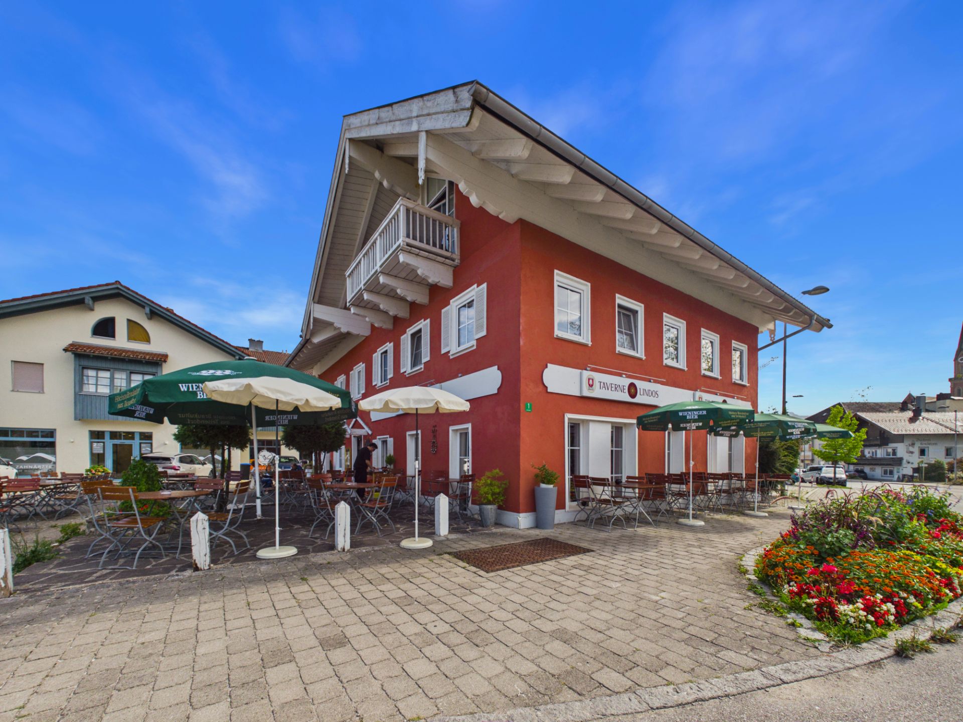 Rotes Wohnhaus mit Balkon und Terrasse, Tische und Sonnenschirmen, Blumenbeet im Vordergrund.