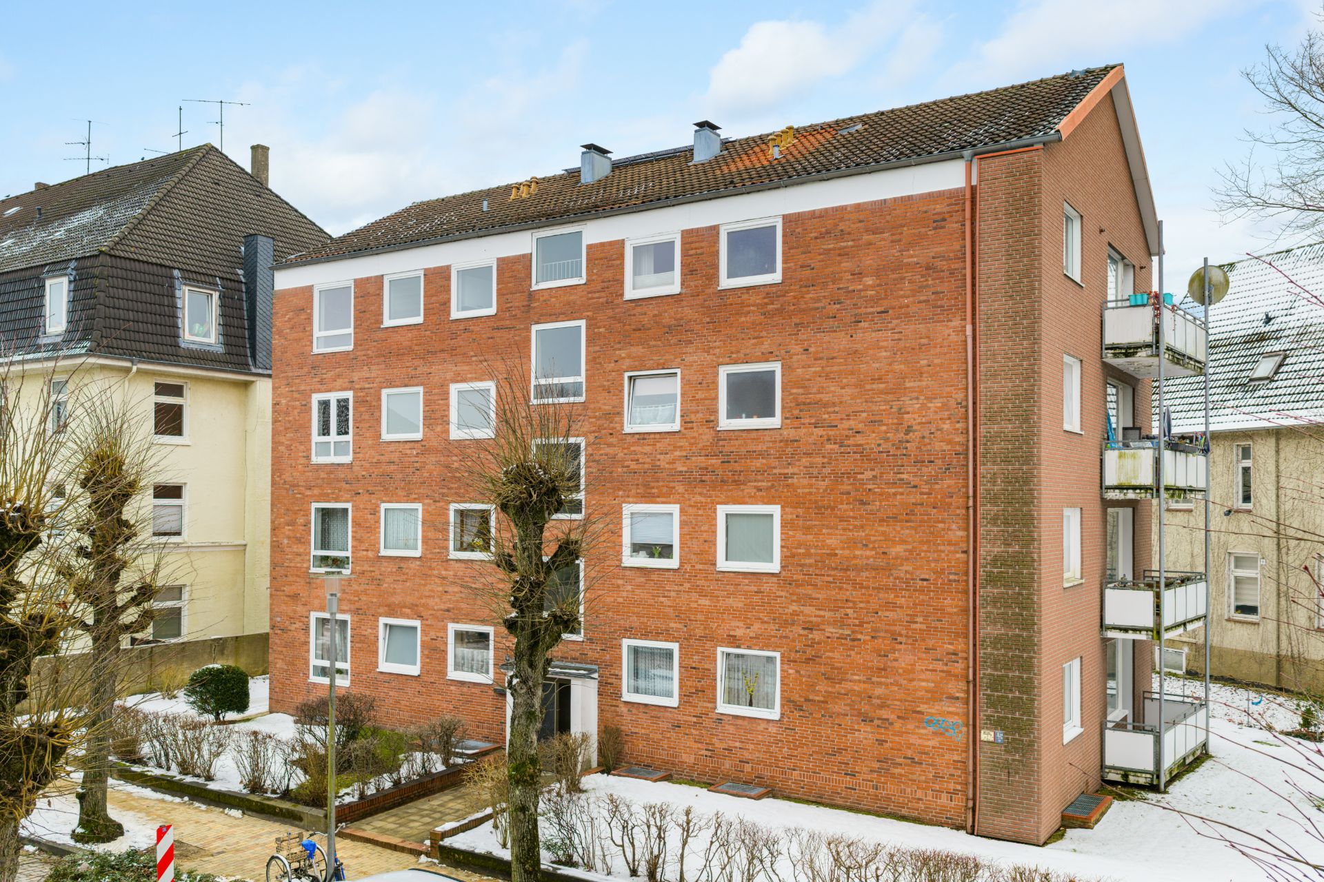 Vierstöckiges Wohnhaus mit roter Klinkerfassade und mehreren Fenstern, Bäume davor, Schnee am Boden.