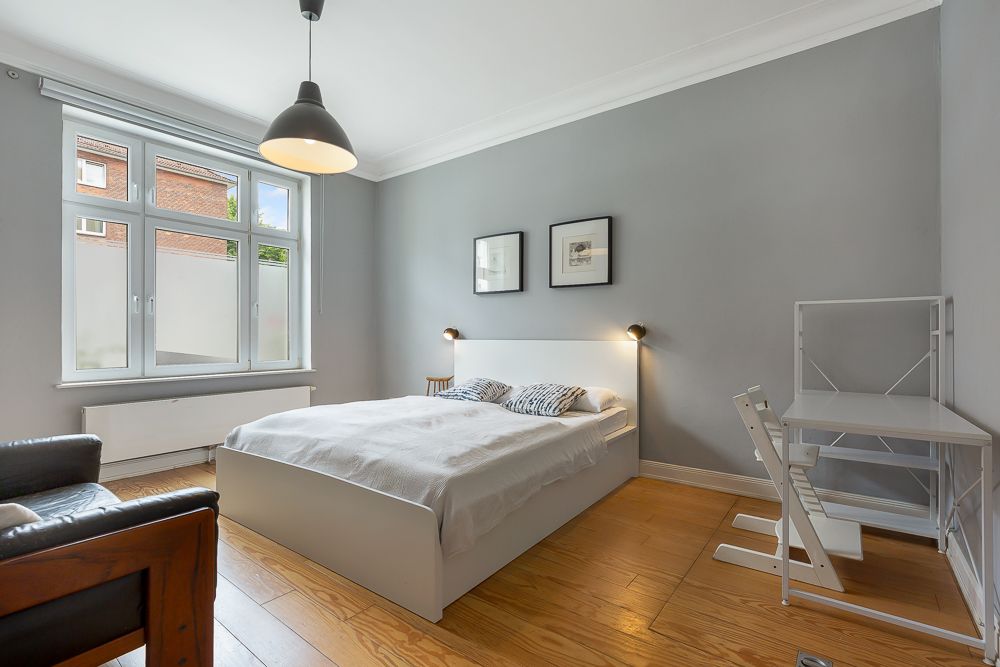 Helles Schlafzimmer mit Doppelbett, Schreibtisch, Stuhl und großem Fenster.