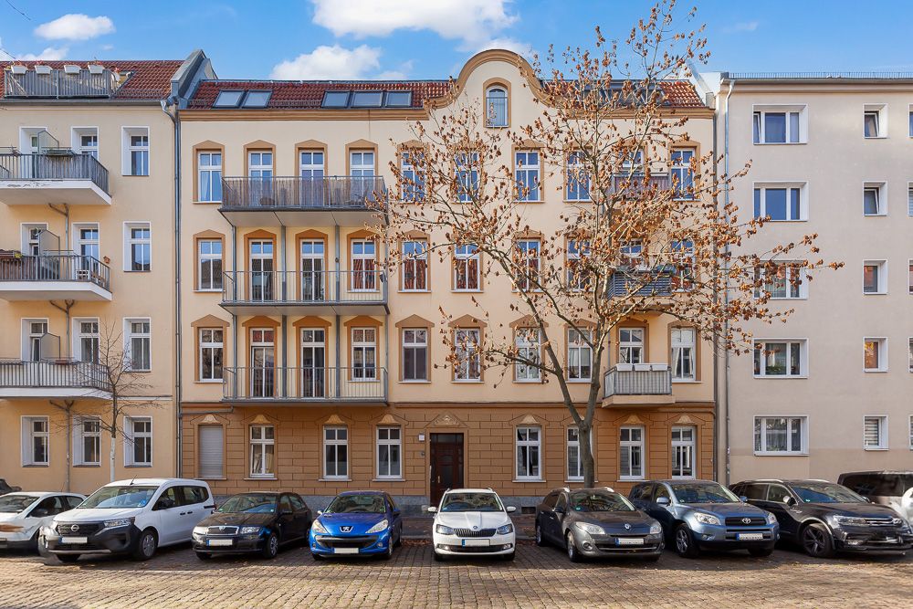 Mehrstöckiges Wohnhaus mit Balkonen, davor parken mehrere Autos und zwei Bäume stehen am Straßenrand.