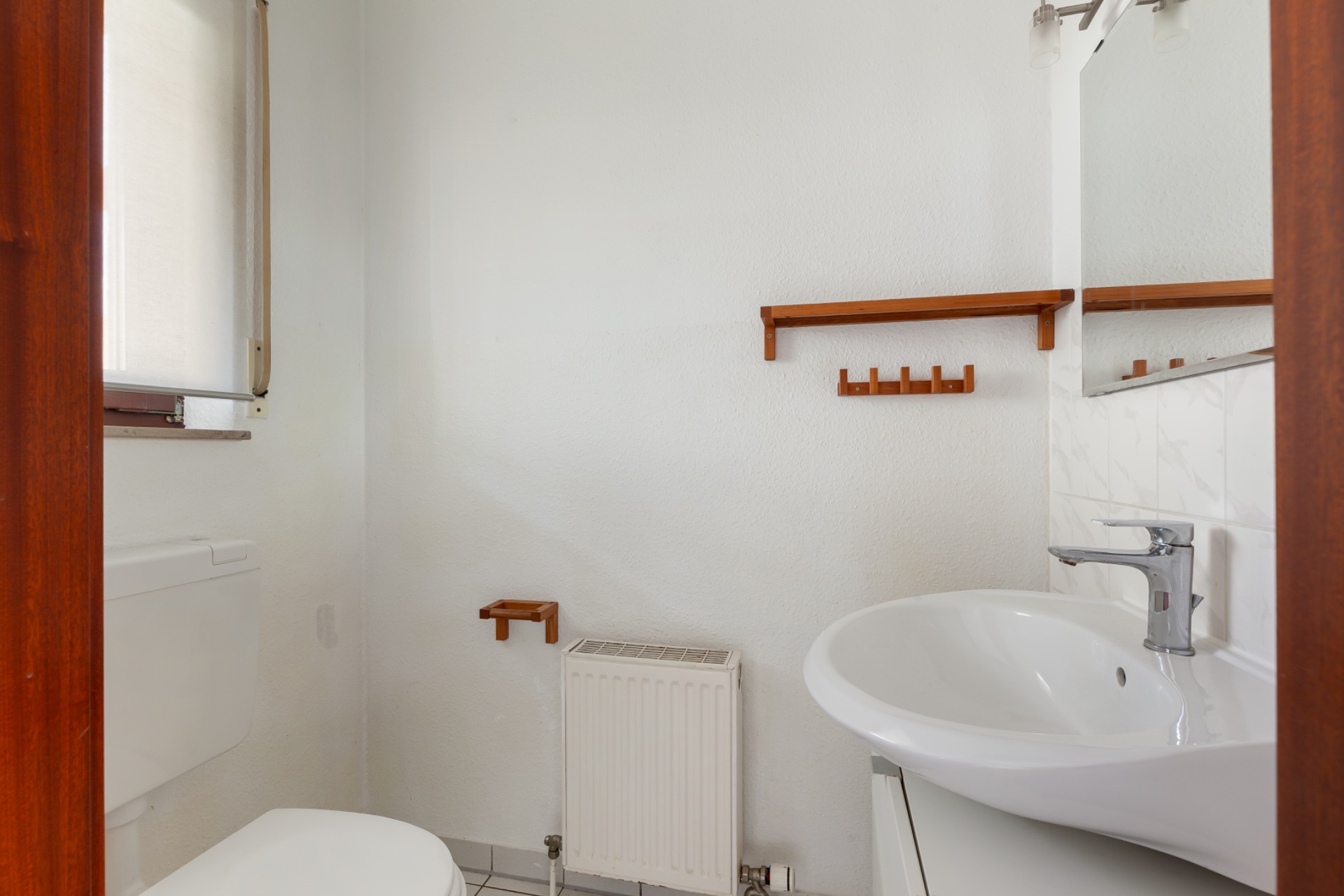 Kleines Badezimmer mit Waschbecken, Toilette, Spiegel und Holzregalen an der Wand.
