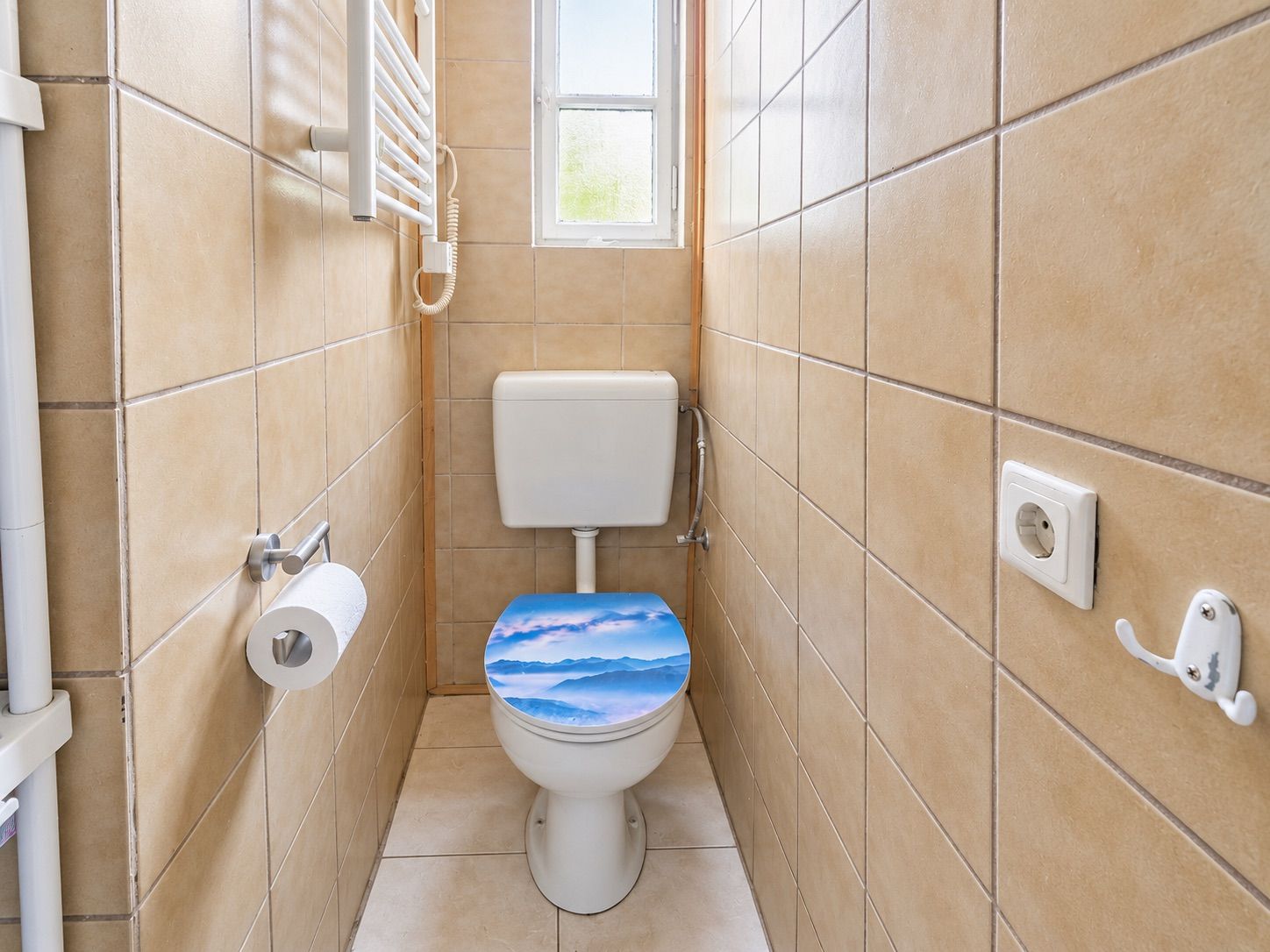 Schmales WC mit beigem Fliesen, Toilettenpapier und Fenster. Toilettendeckel mit Landschaftsmotiv.
