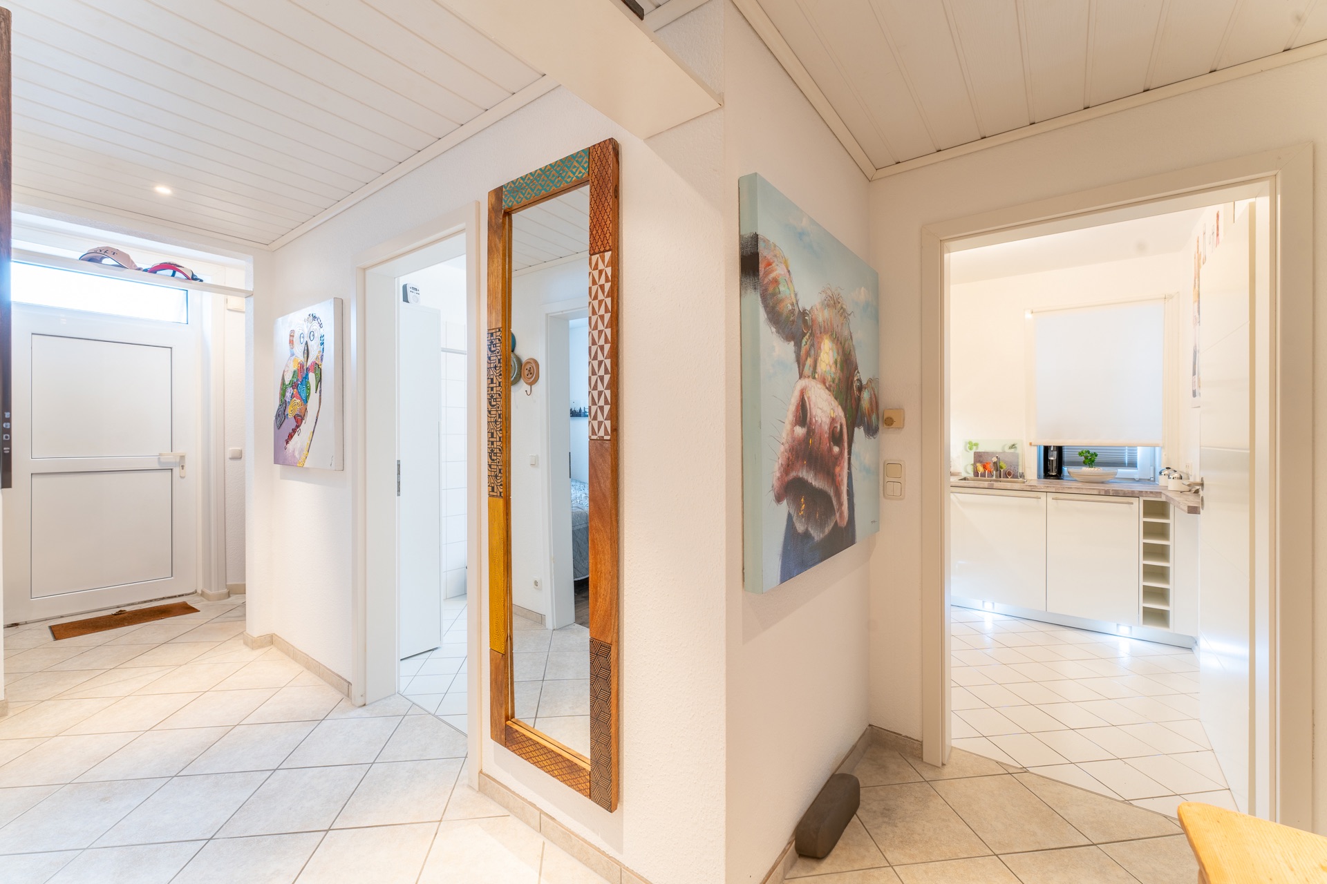 Heller Flur mit Bodenfliesen, großem Spiegel, bunter Kunst und Blick in die Küche.