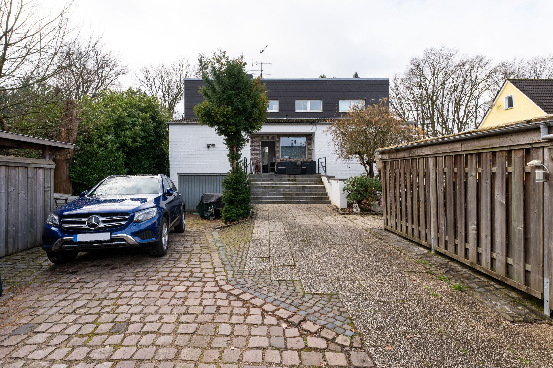 Vor einem weißen Wohnhaus steht ein blauer SUV auf einer Einfahrt mit Pflastersteinen.