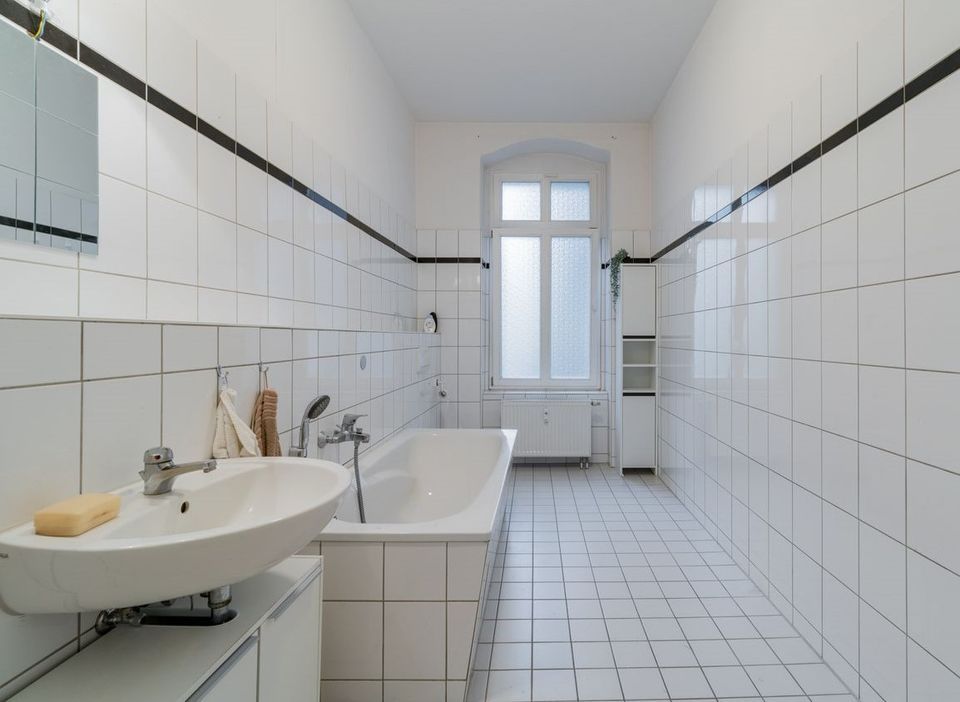 Helles Badezimmer mit Badewanne, Waschbecken und großem Fenster, weiße Fliesen an Wänden und Boden.