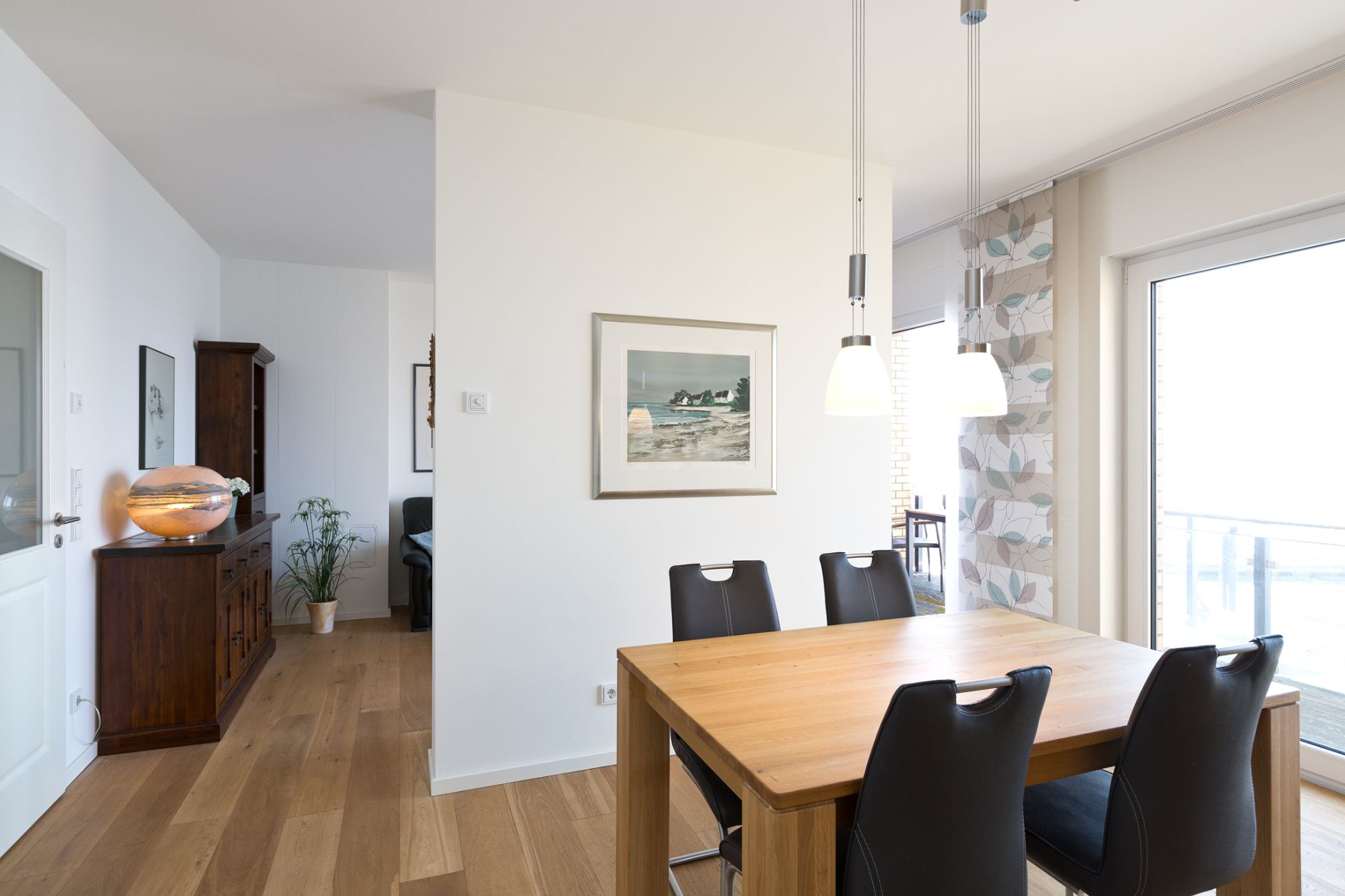 Essbereich mit Holztisch und vier schwarzen Stühlen, helle Wände, große Fenster, hängende Lampen.