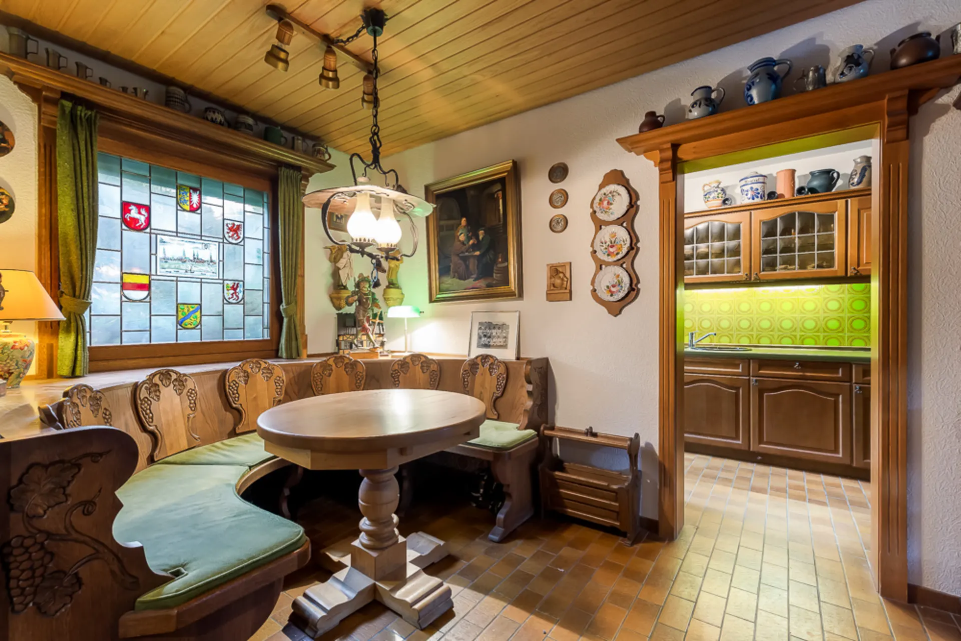 Gemütliche Essecke mit Holztisch, Eckbank, buntem Fenster und Blick in Holz-Küche mit grüner Fliesenwand.