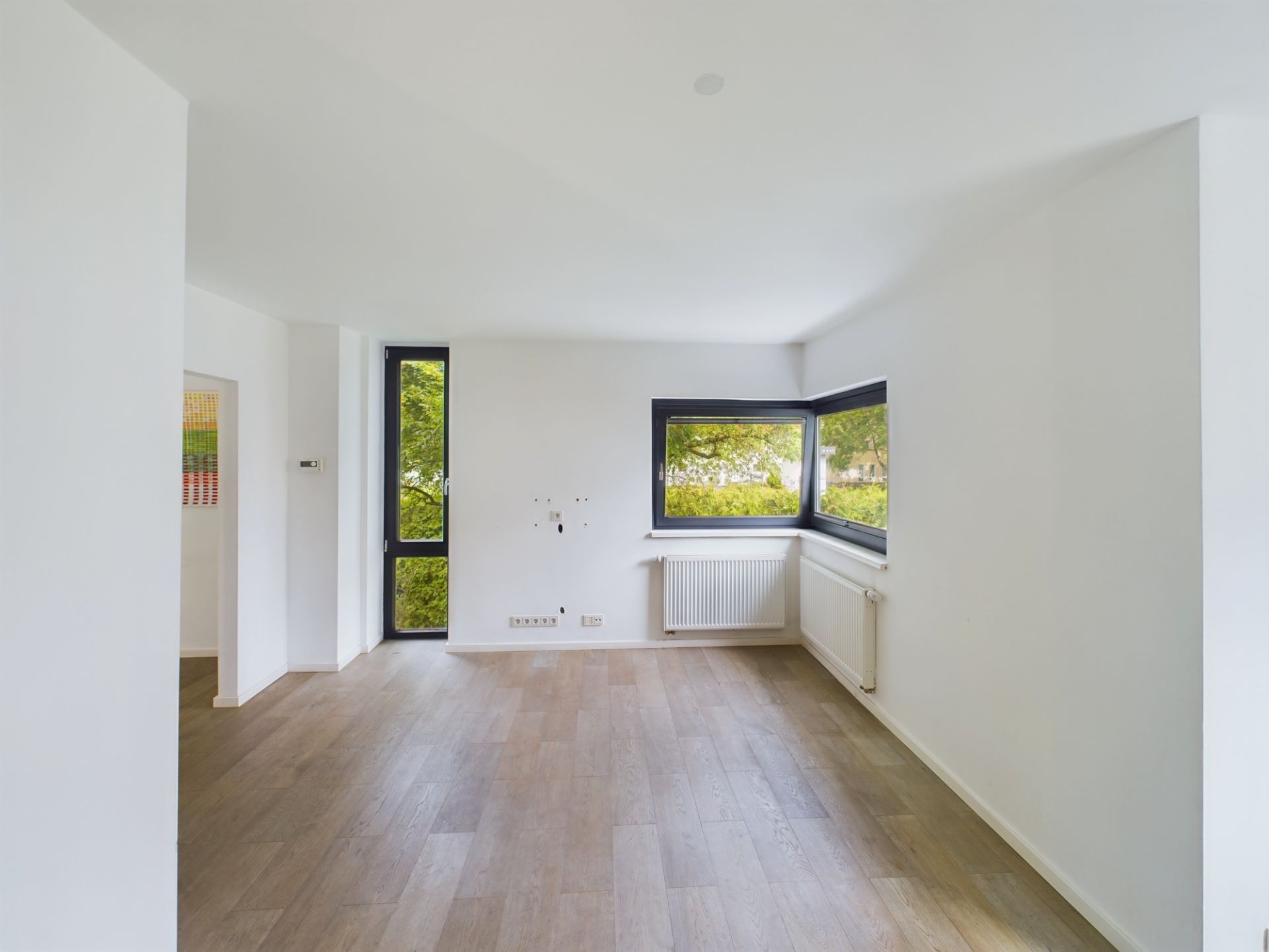 Leerer Raum mit Holzfußboden, weißen Wänden und großen Fenstern mit Blick ins Grüne.