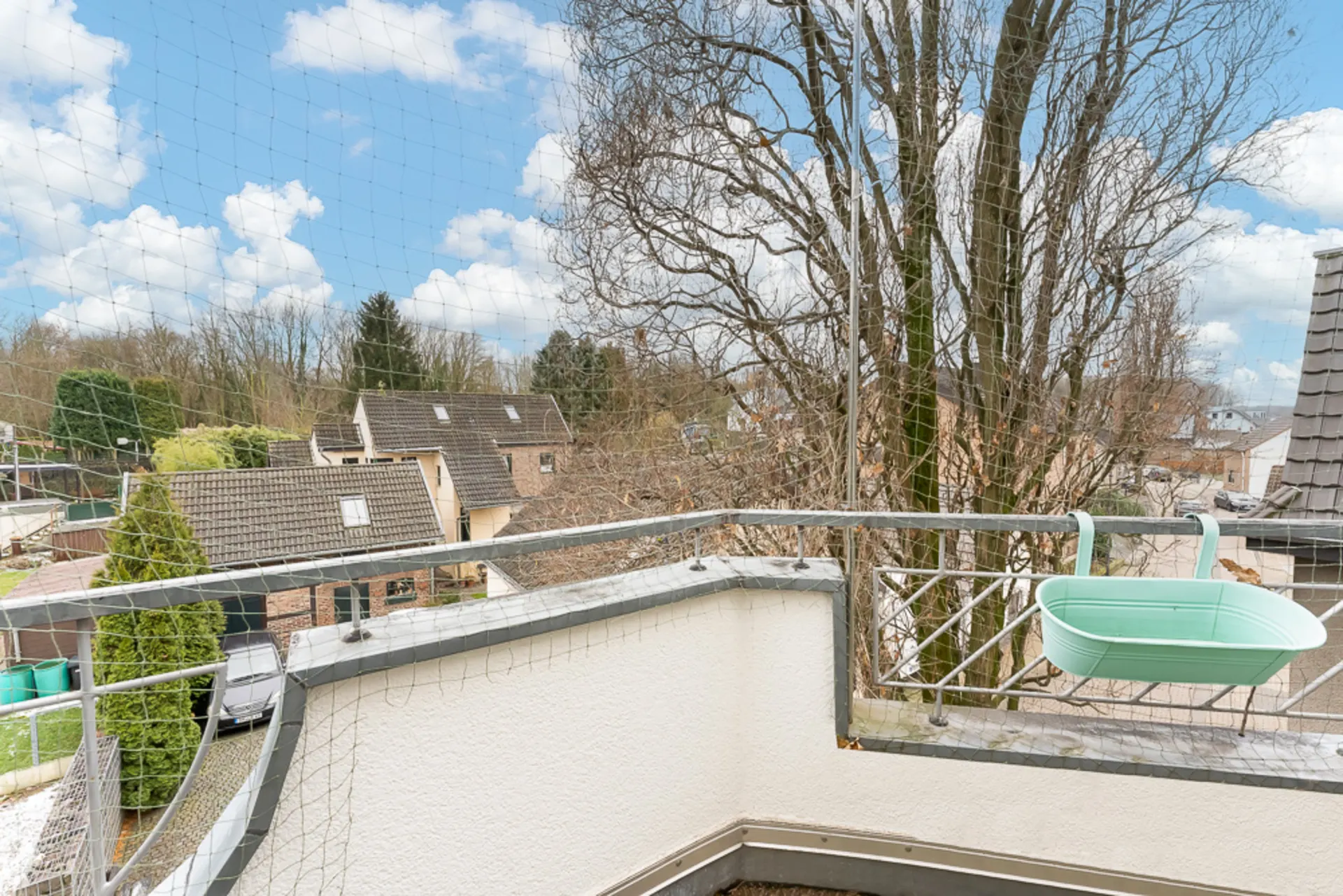 Balkon mit Metallgeländer und grünem Blumenkasten, Blick auf Bäume und Nachbarhäuser.
