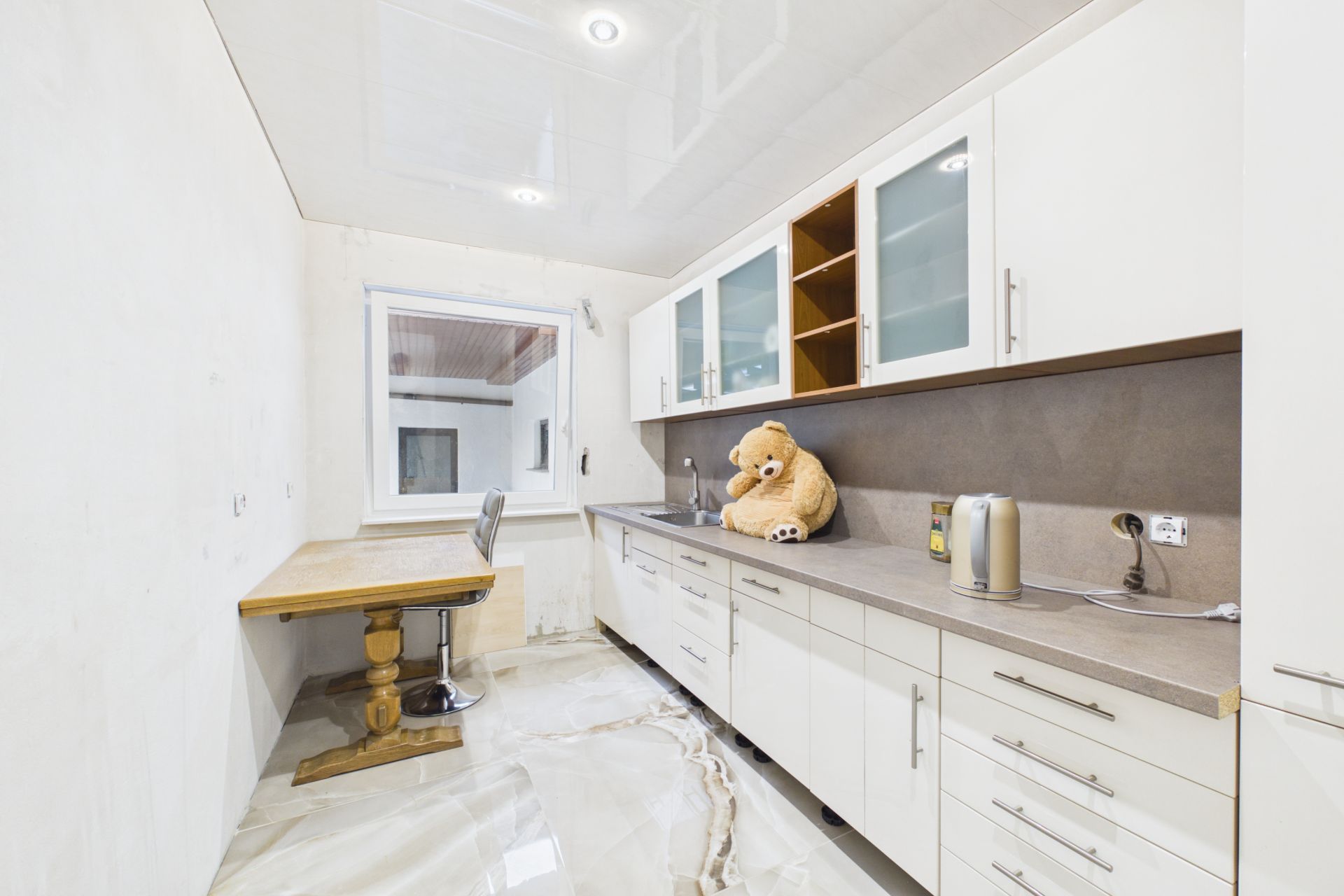 Helle Küche mit weißen Schränken, Fenster, Schreibtisch, Stuhl und großem Teddybär auf der Arbeitsplatte.