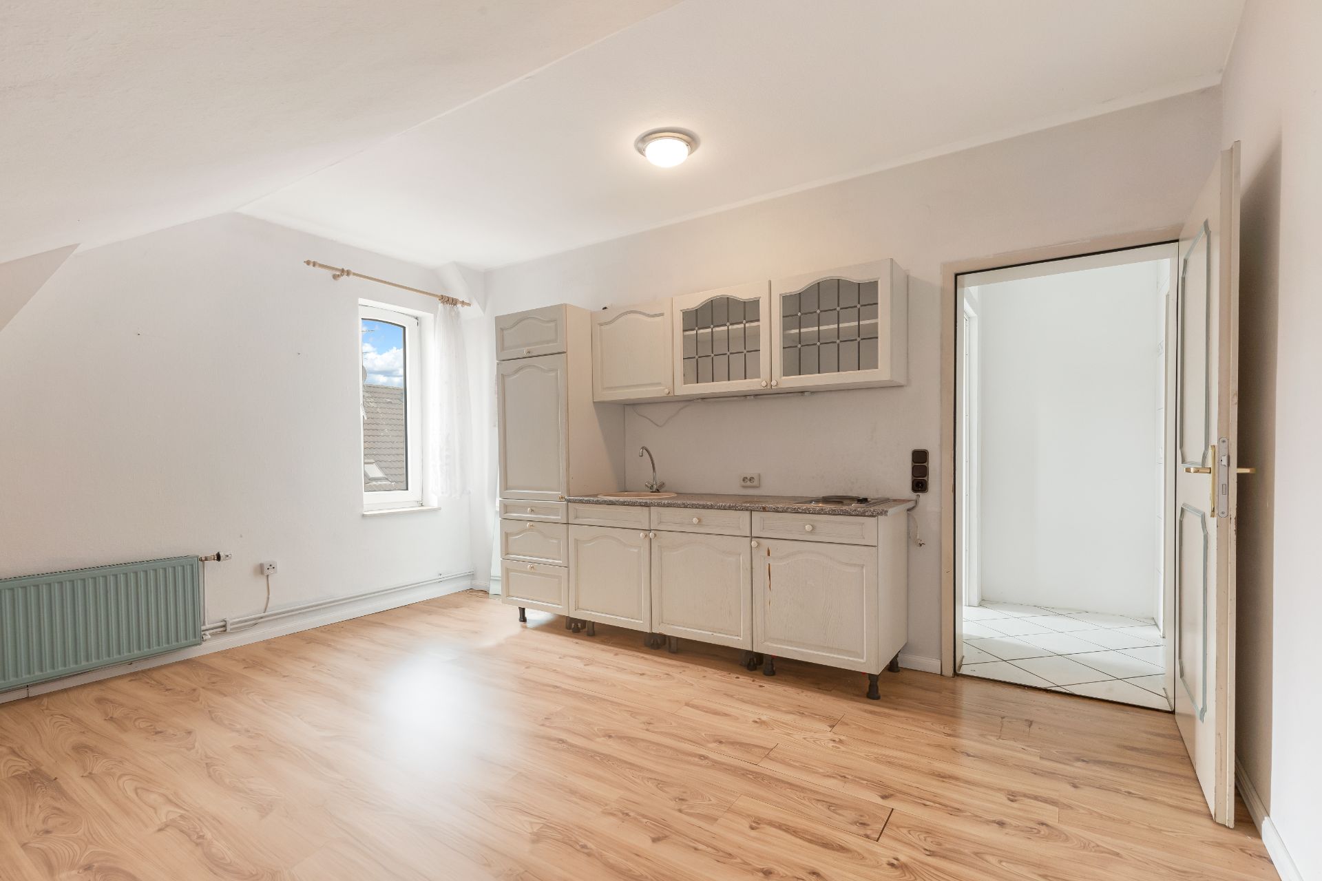 Heller Raum mit Holzboden, kleiner Einbauküche, Fenster und offenem Durchgang zu einem weiteren Zimmer.