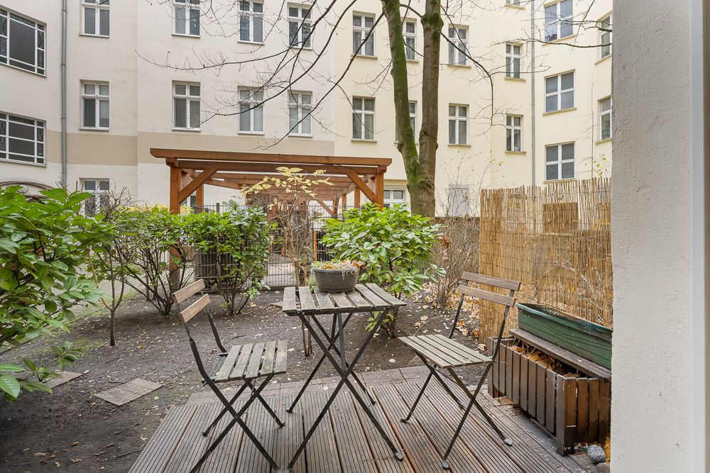 Kleiner Garten mit Holztisch, drei Stühlen, Pflanzen und Blick auf ein Wohnhaus.