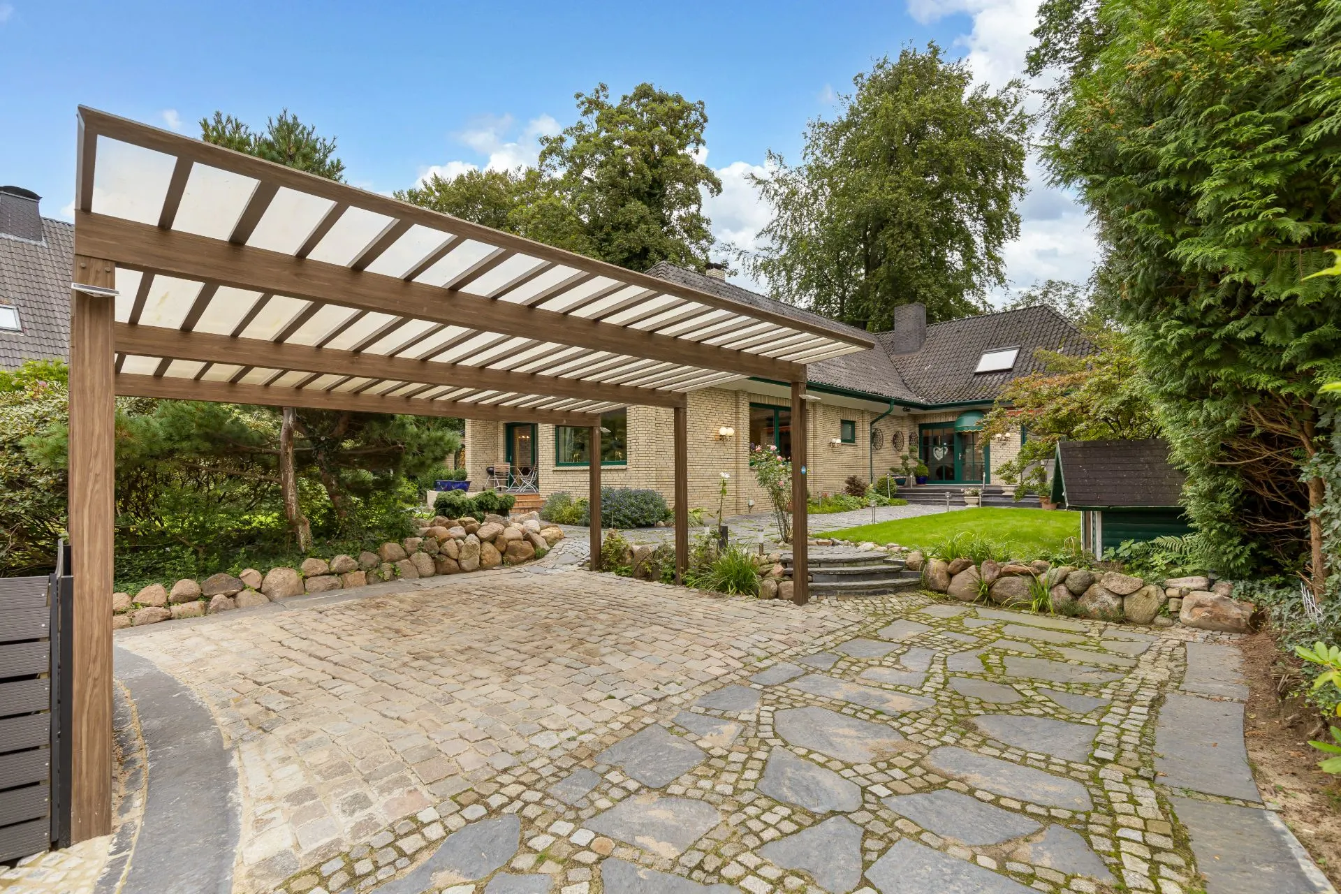 Einfahrt mit Carport, Kopfsteinpflaster und Blick auf ein Wohnhaus mit Garten.