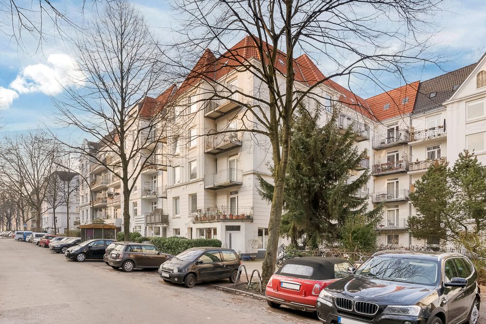 Mehrstöckiges Wohnhaus mit Balkonen, Bäumen und parkenden Autos an der Straße.