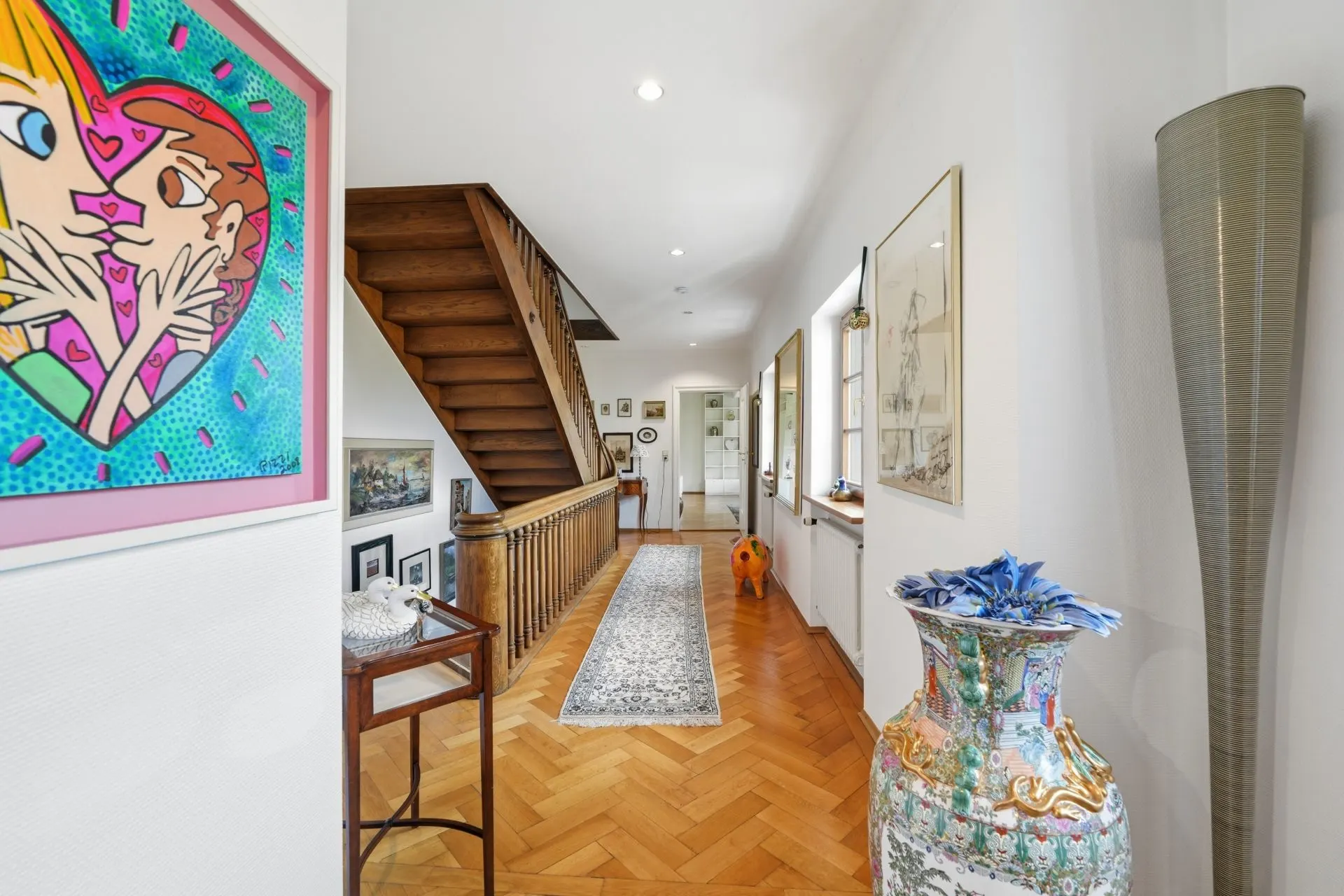Heller Flur mit Holztreppe, Teppichläufer, bunter Vase und Wandbildern in einem Wohnhaus.