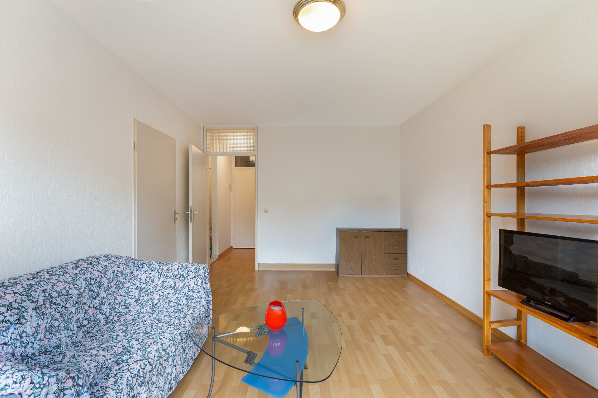Wohnzimmer mit gemustertem Sofa, Glastisch, Fernseherregal und Kommode auf hellem Holzboden.