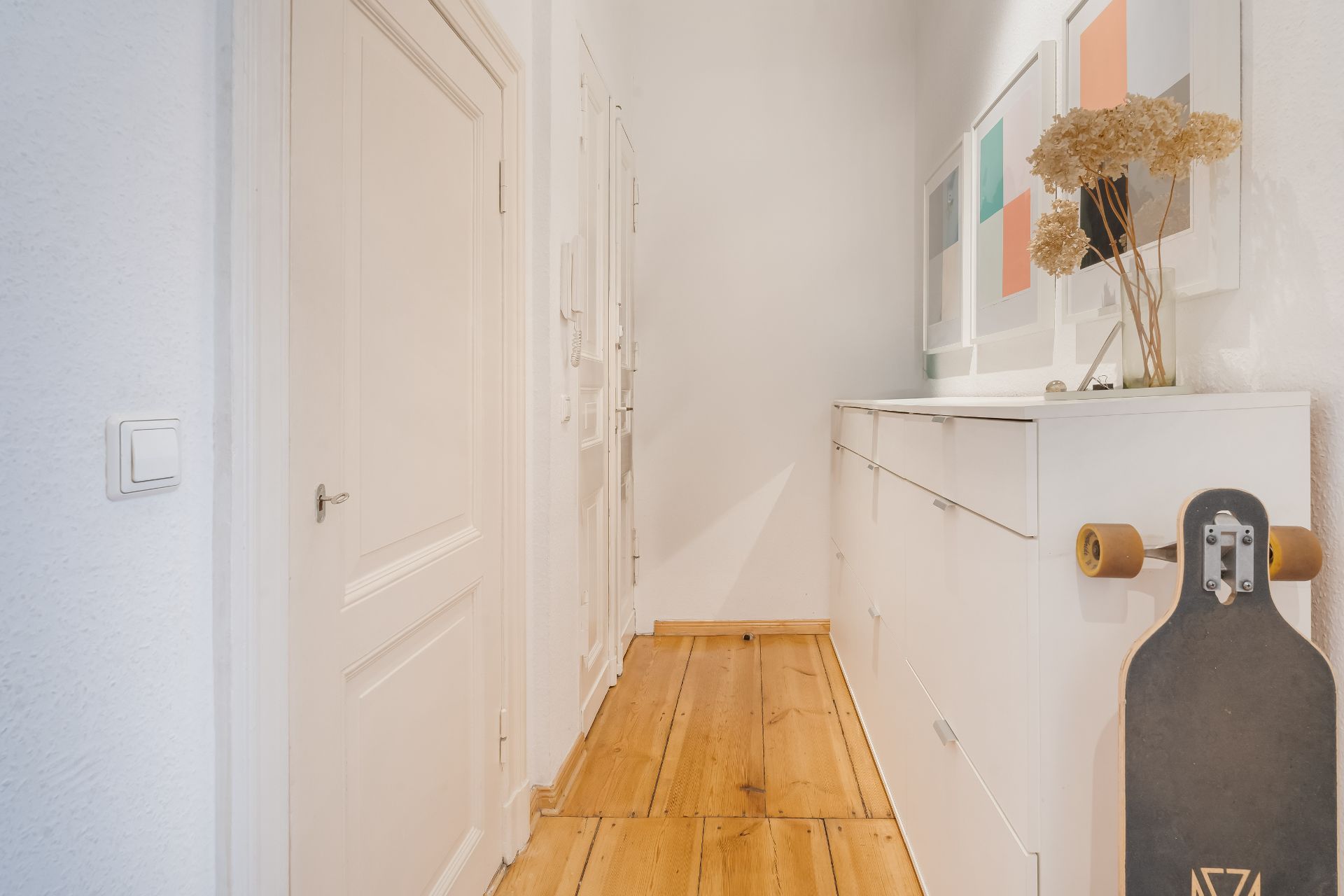 Heller Flur mit Holzboden, weißem Sideboard, Bilderrahmen und Longboard an der Wand.
