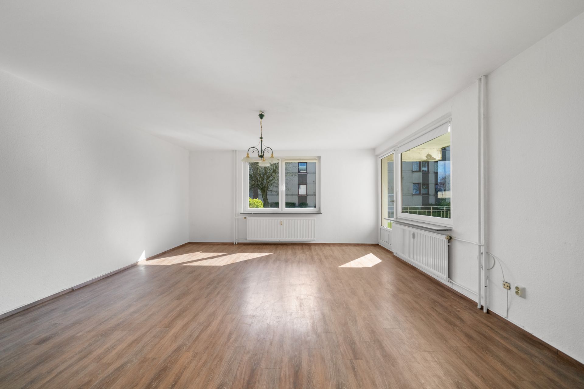 Helles, leeres Zimmer mit Holzfußboden, weißen Wänden und zwei großen Fenstern.