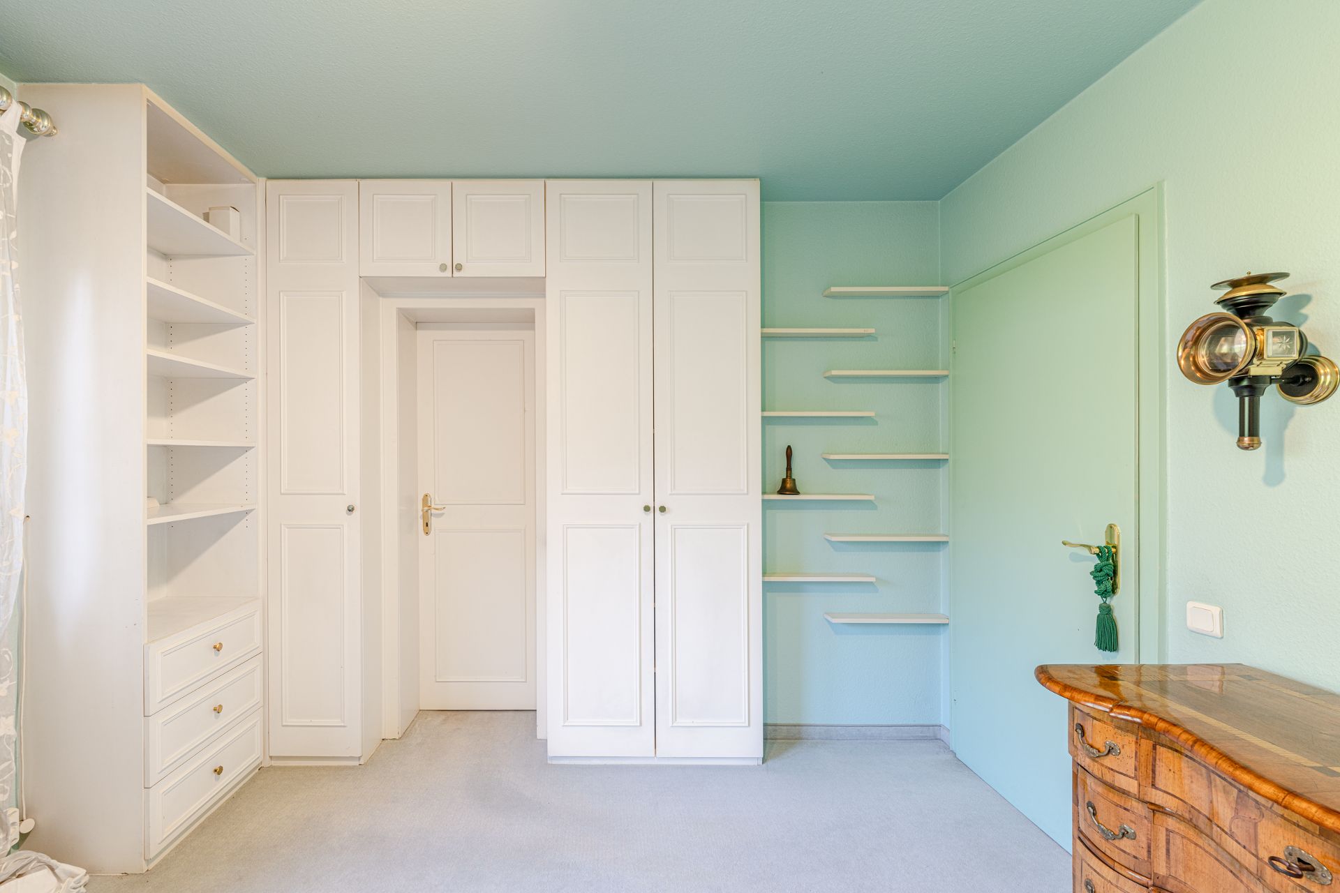 Helles Zimmer mit weißen Einbauschränken, offenen Regalen und einer Kommode aus Holz.