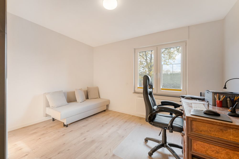 Helles Zimmer mit Holzfußboden, Sofa, Schreibtisch, Bürostuhl und zwei Fenstern.