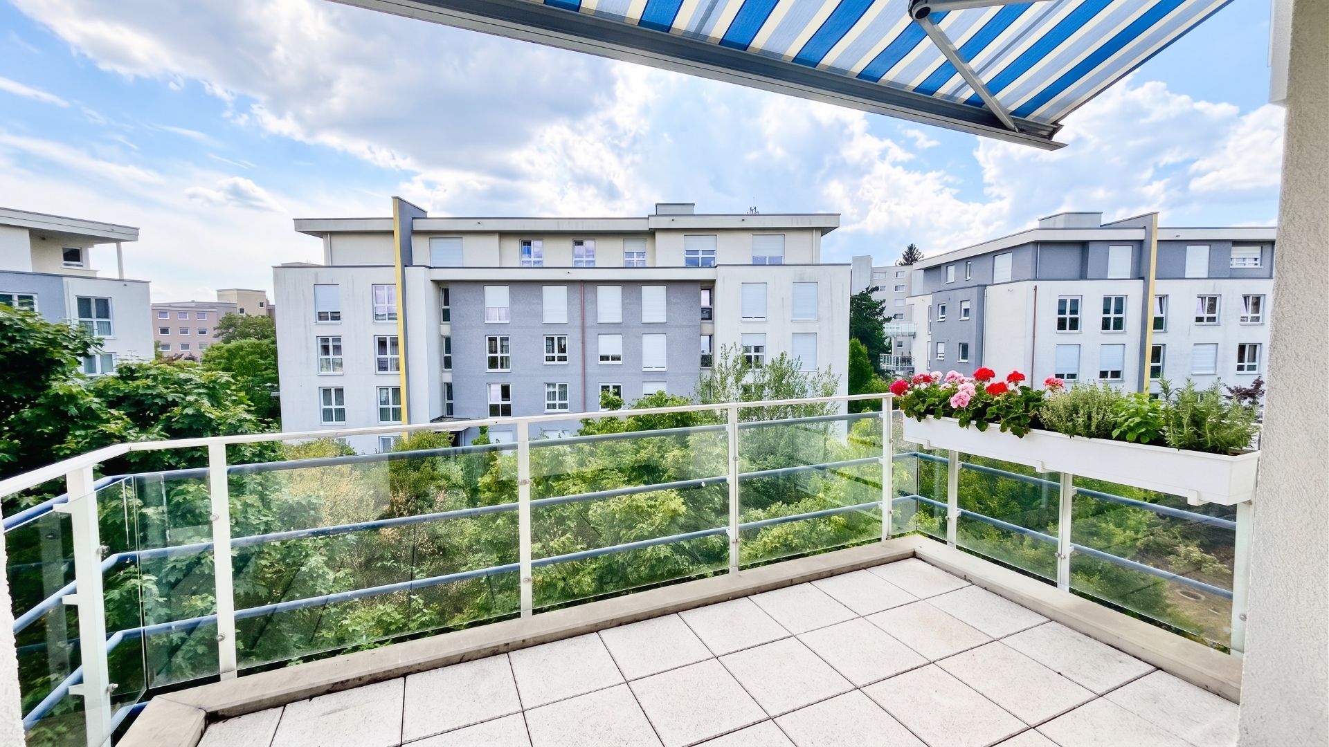 Balkon mit Glasgeländer, Markise und Blumenkasten, Blick auf Wohnhäuser und Bäume.