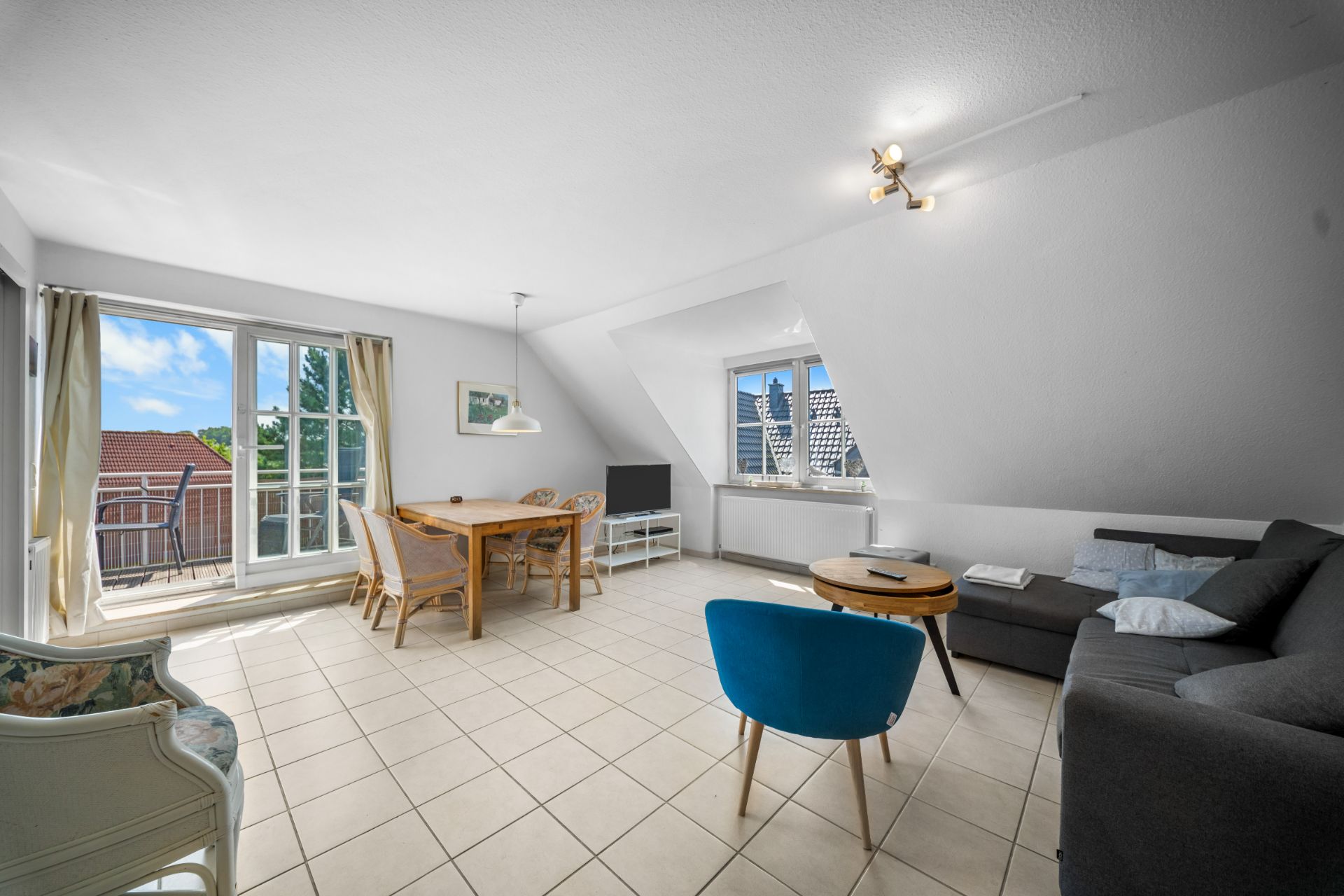 Helles Wohnzimmer mit Sofa, Esstisch, Stühlen, Fernseher und Zugang zum Balkon.