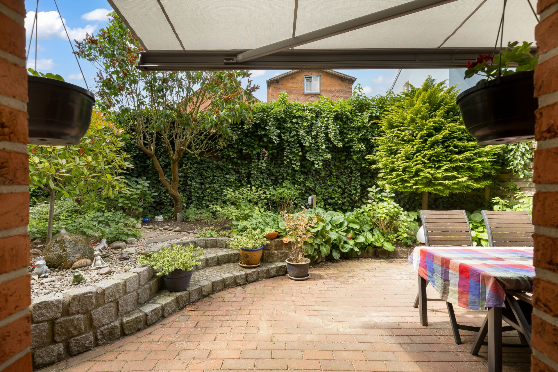 Überdachte Terrasse mit Tisch und Stühlen, umgeben von bepflanztem Garten und Steinmauer.