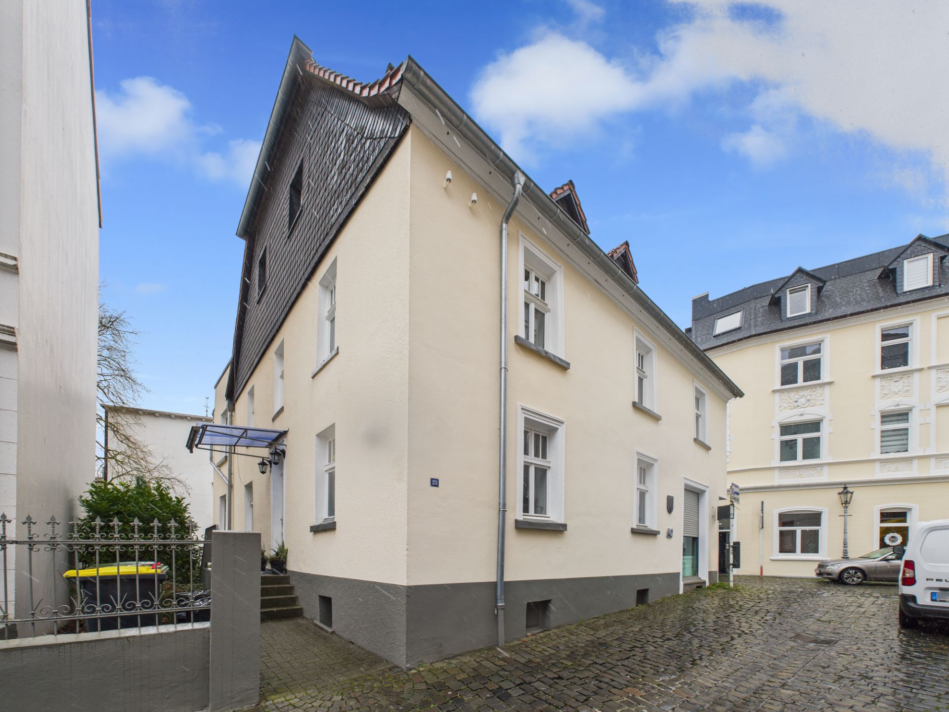 Zweistöckiges, helles Wohnhaus mit dunklem Dach und mehreren Fenstern auf gepflastertem Hof.