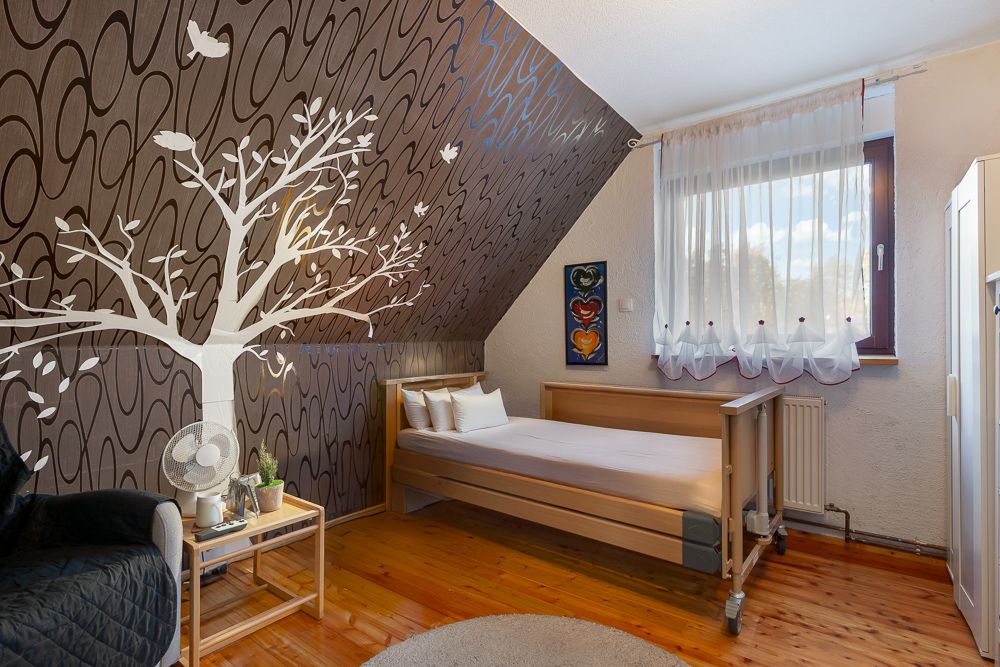 Helles Schlafzimmer mit Einzelbett, Schrägdecke, Wandtapete mit Baum und großem Fenster.