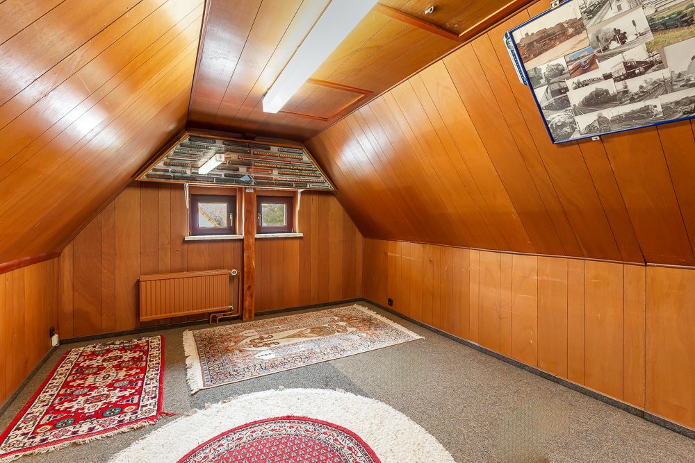 Dachzimmer mit Holzvertäfelung, Teppichen, zwei Fenstern und Heizung an der Stirnwand.