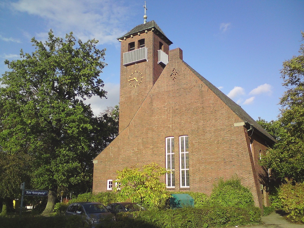 Backsteingebäude mit hohem Turm und Uhr, umgeben von Bäumen und Büschen.