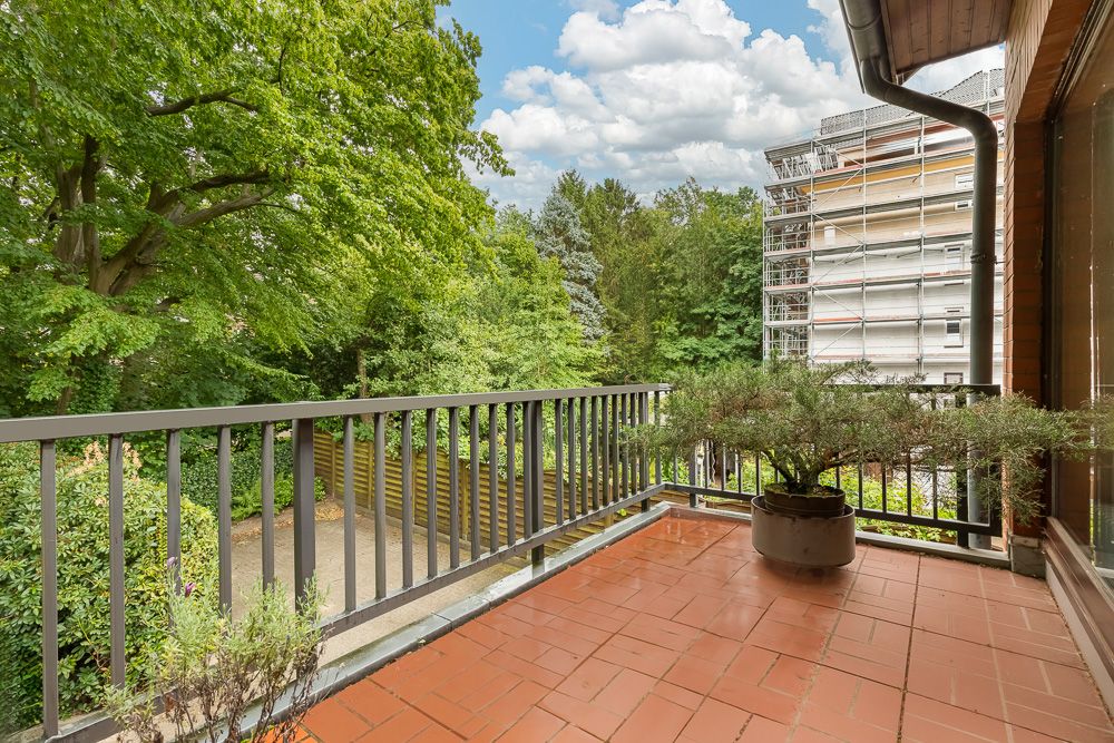 Balkon mit rot gefliestem Boden, Geländer, Pflanzen und Blick auf Bäume und ein Baugerüst.