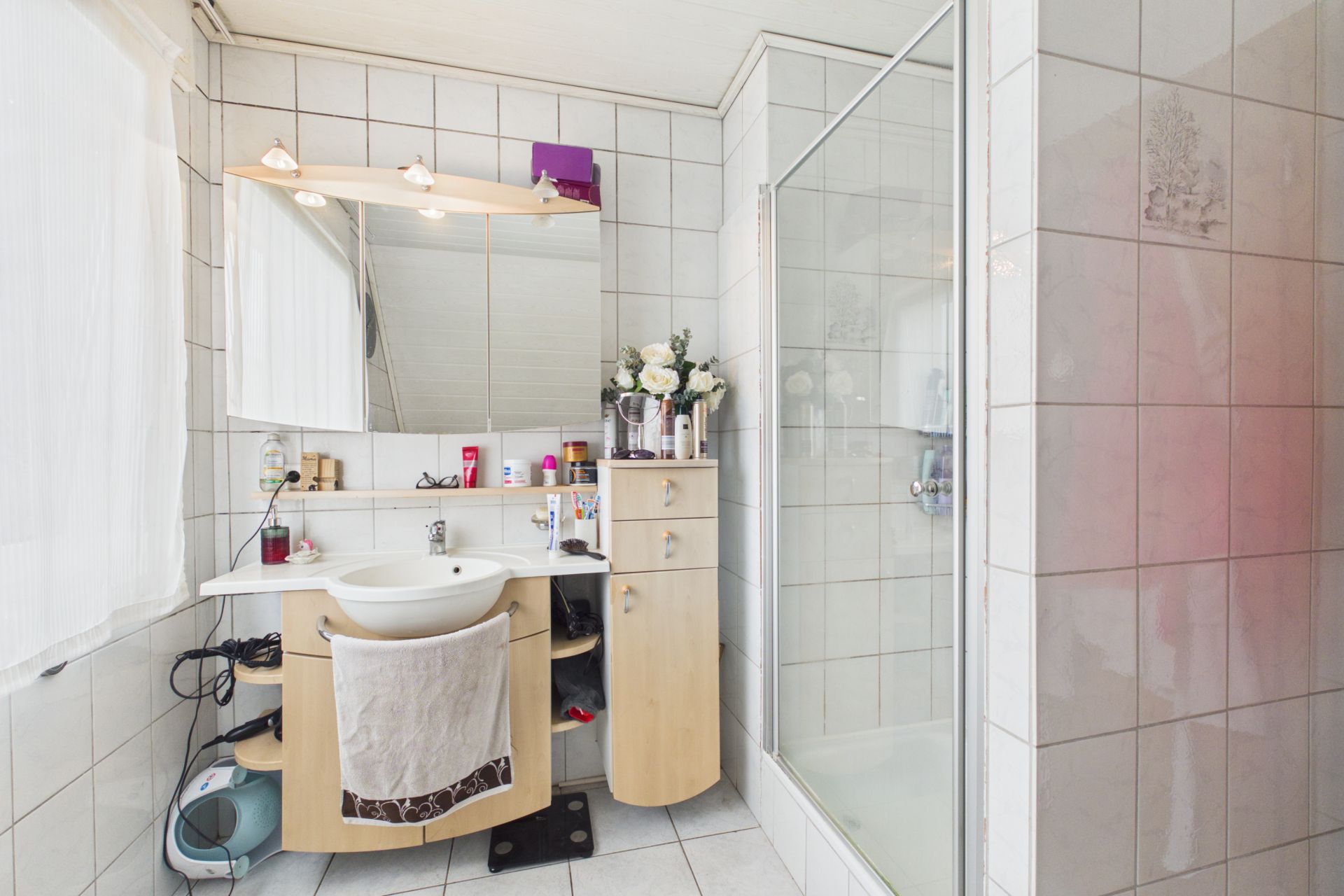 Helles Badezimmer mit Dusche, Waschbecken, Spiegel und Holzschrank auf weißen Fliesen.