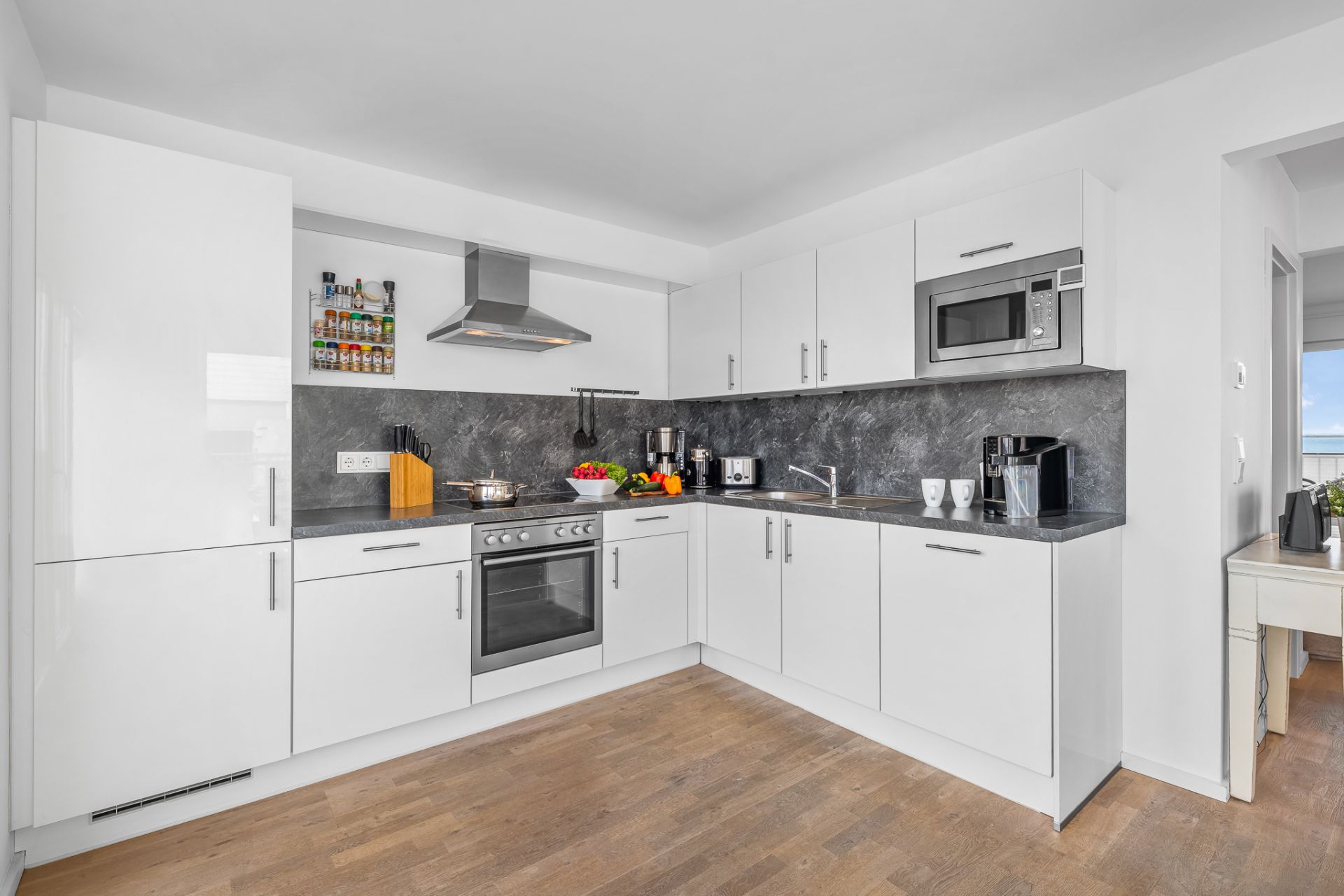 Moderne weiße Einbauküche mit Geräten, grauer Arbeitsplatte und Holzfußboden.