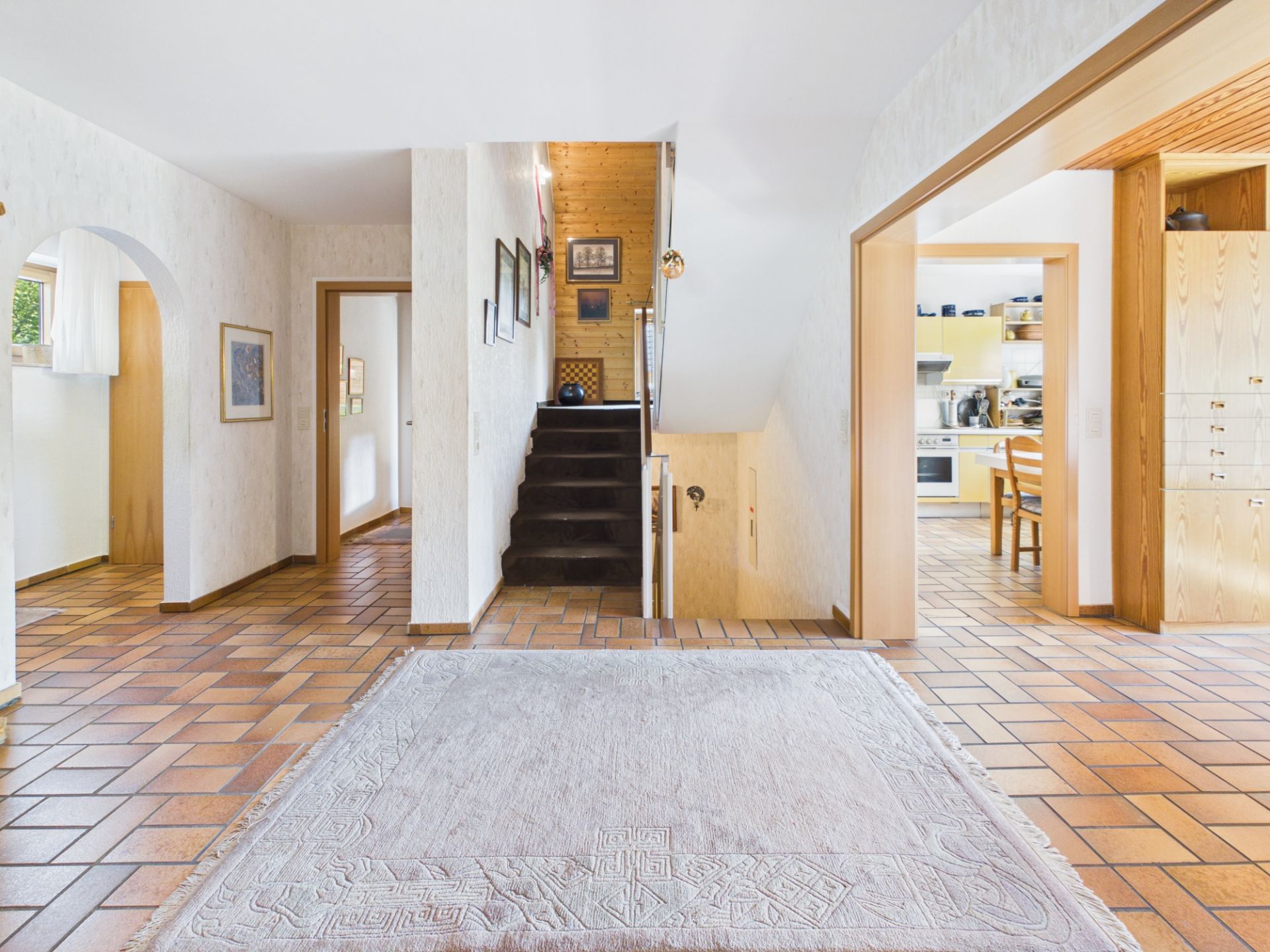 Heller Flur mit Teppich, braunen Fliesen, Treppe und angrenzender Küche in einem Wohnhaus.