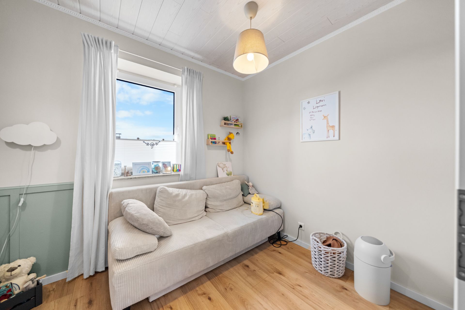 Helles Kinderzimmer mit Sofa, Spielzeugkorb, Wandregal und Fenster mit weißen Vorhängen.