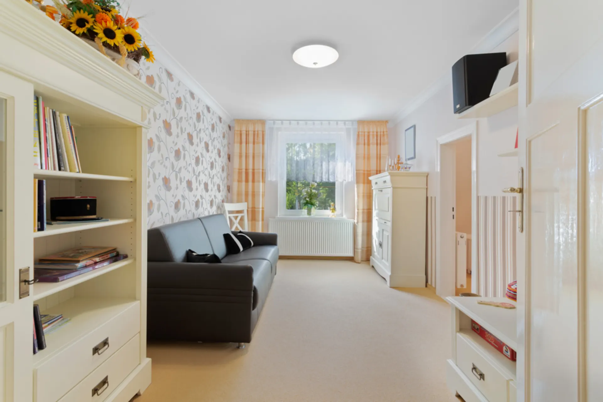 Wohnzimmer mit schwarzem Sofa, Bücherregal, Kommode und Fenster mit Vorhängen.