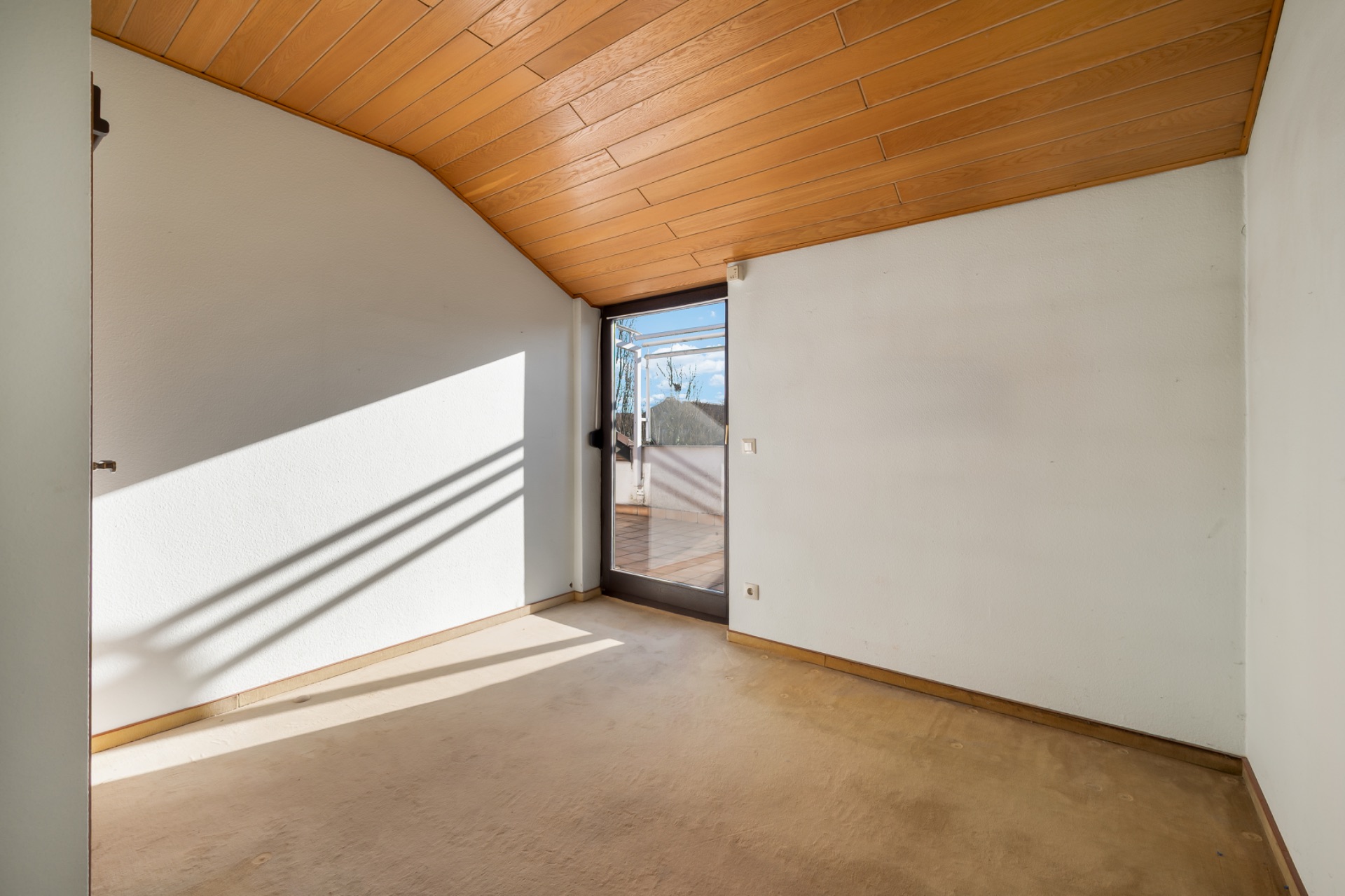 Leerer Raum mit schräger Holzdecke und Glastür zu einem Balkon.