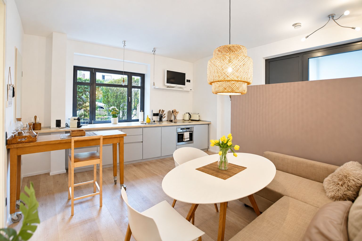 Helle Wohnküche mit Esstisch, Sofa, Küchenzeile und großen Fenstern, dekoriert mit Blumen.