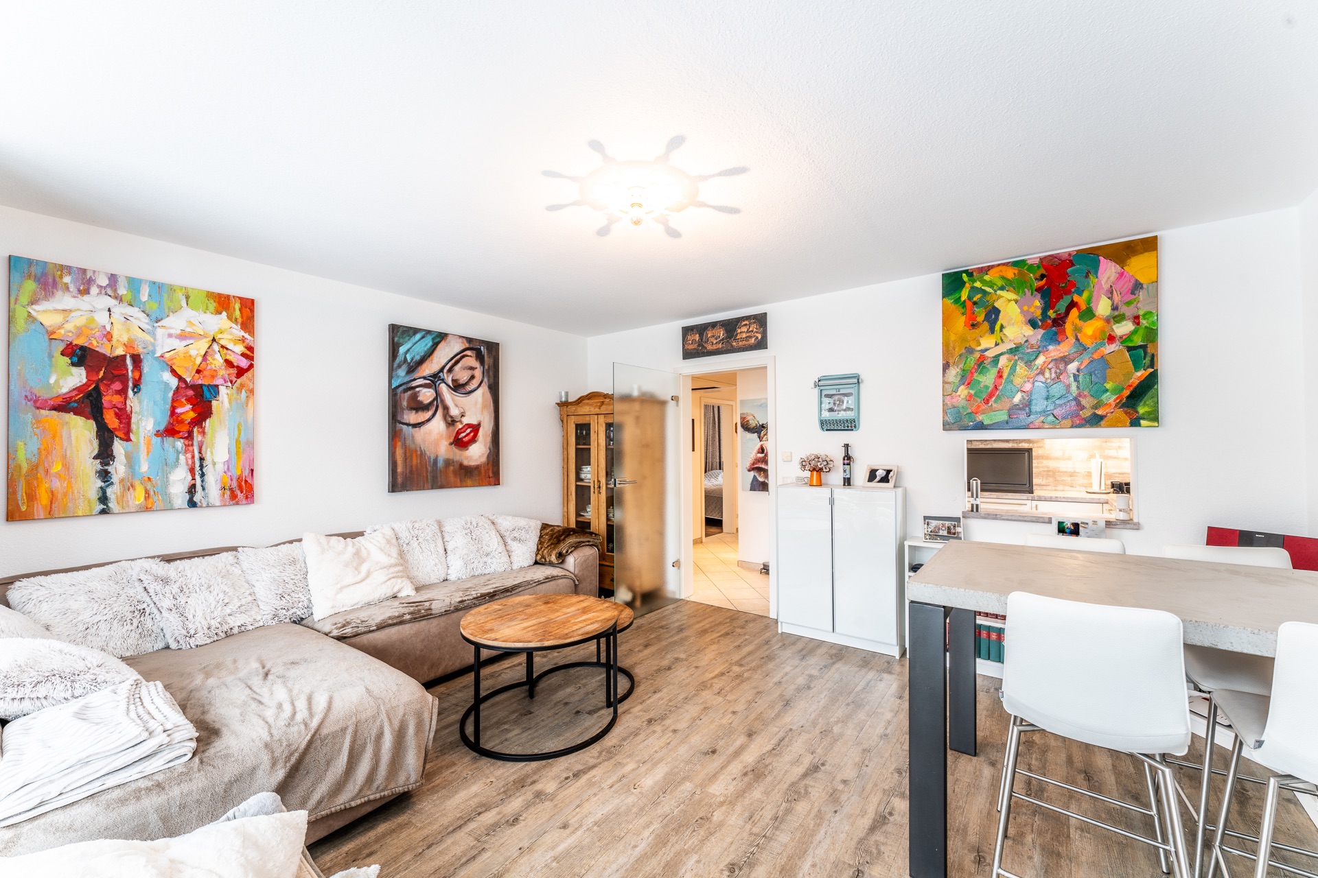 Helles Wohnzimmer mit Ecksofa, Couchtisch, Barhockern und bunten Gemälden an den Wänden.