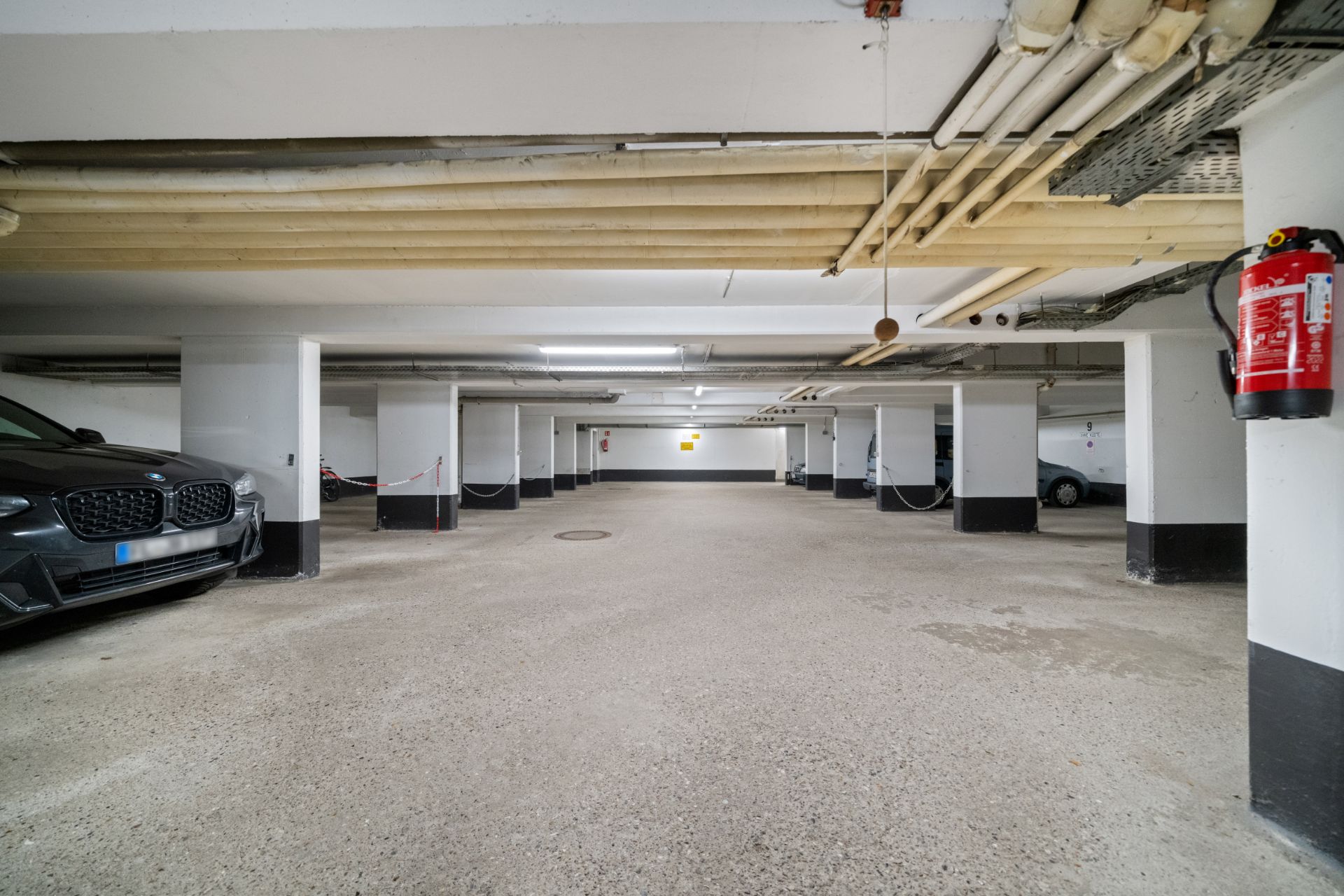 Tiefgarage mit geparkten Autos, weißen Wänden und sichtbaren Rohren an der Decke.