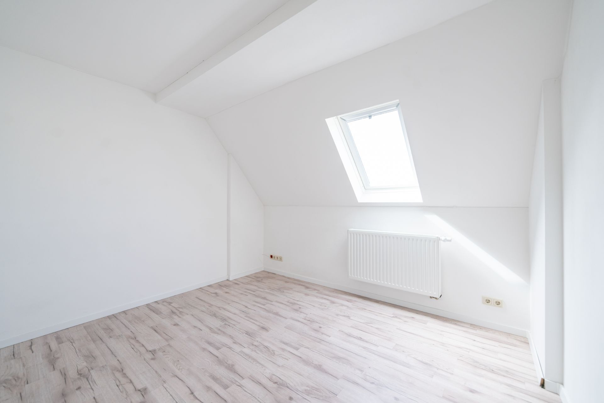 Helles, leeres Zimmer mit schräger Decke, Dachfenster und hellen Holzboden.