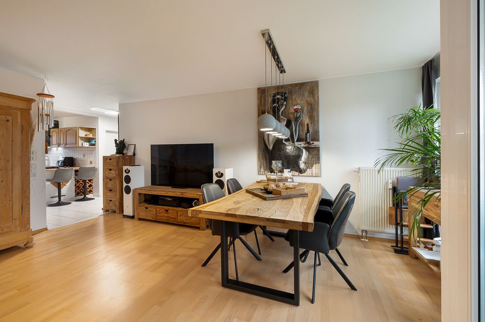 Wohnzimmer mit Holztisch, schwarzen Stühlen, großem Fernseher und Pflanze am Fenster.