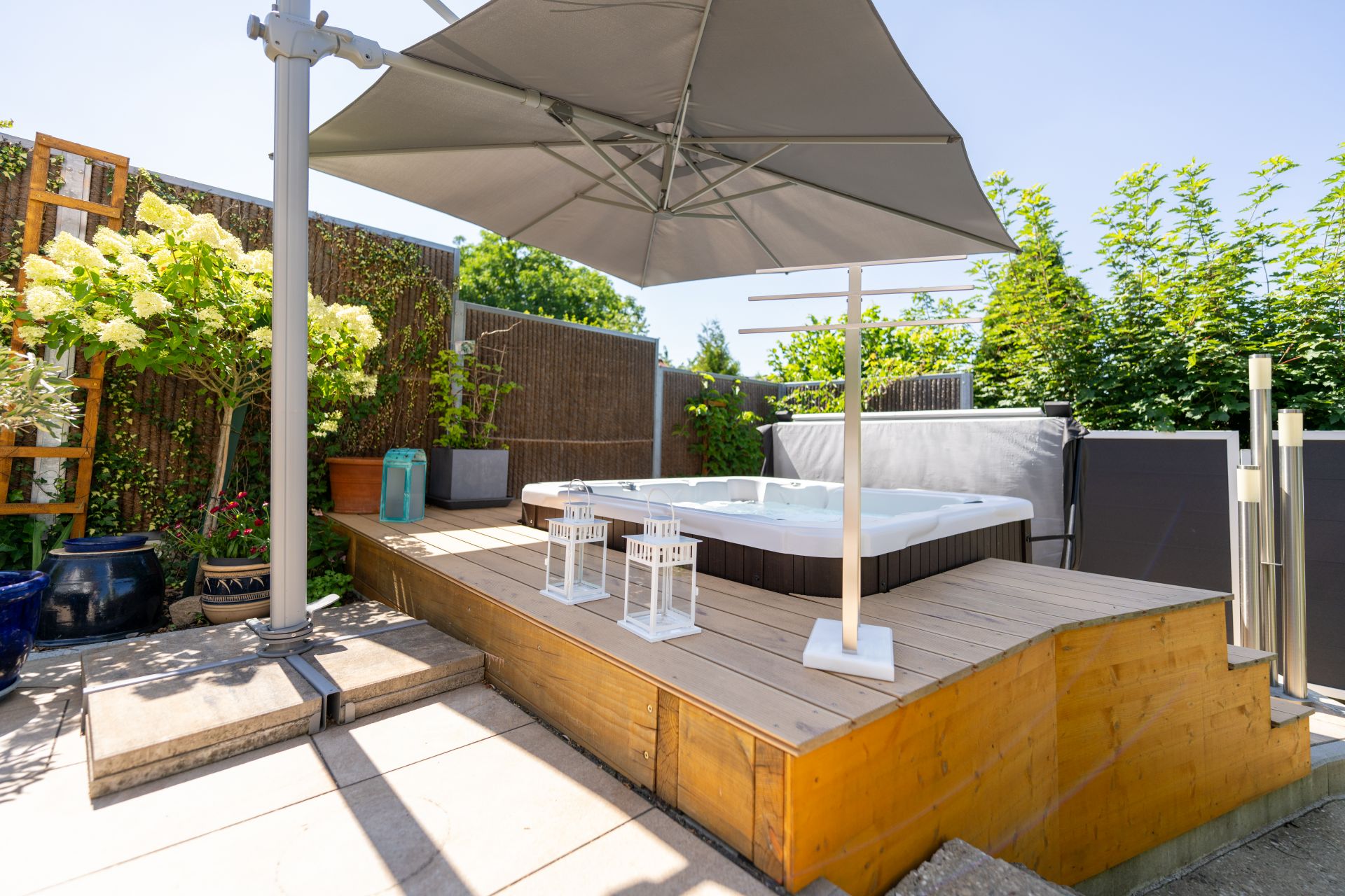 Holzterrasse mit Whirlpool, Sonnenschirm und Pflanzen im Garten.