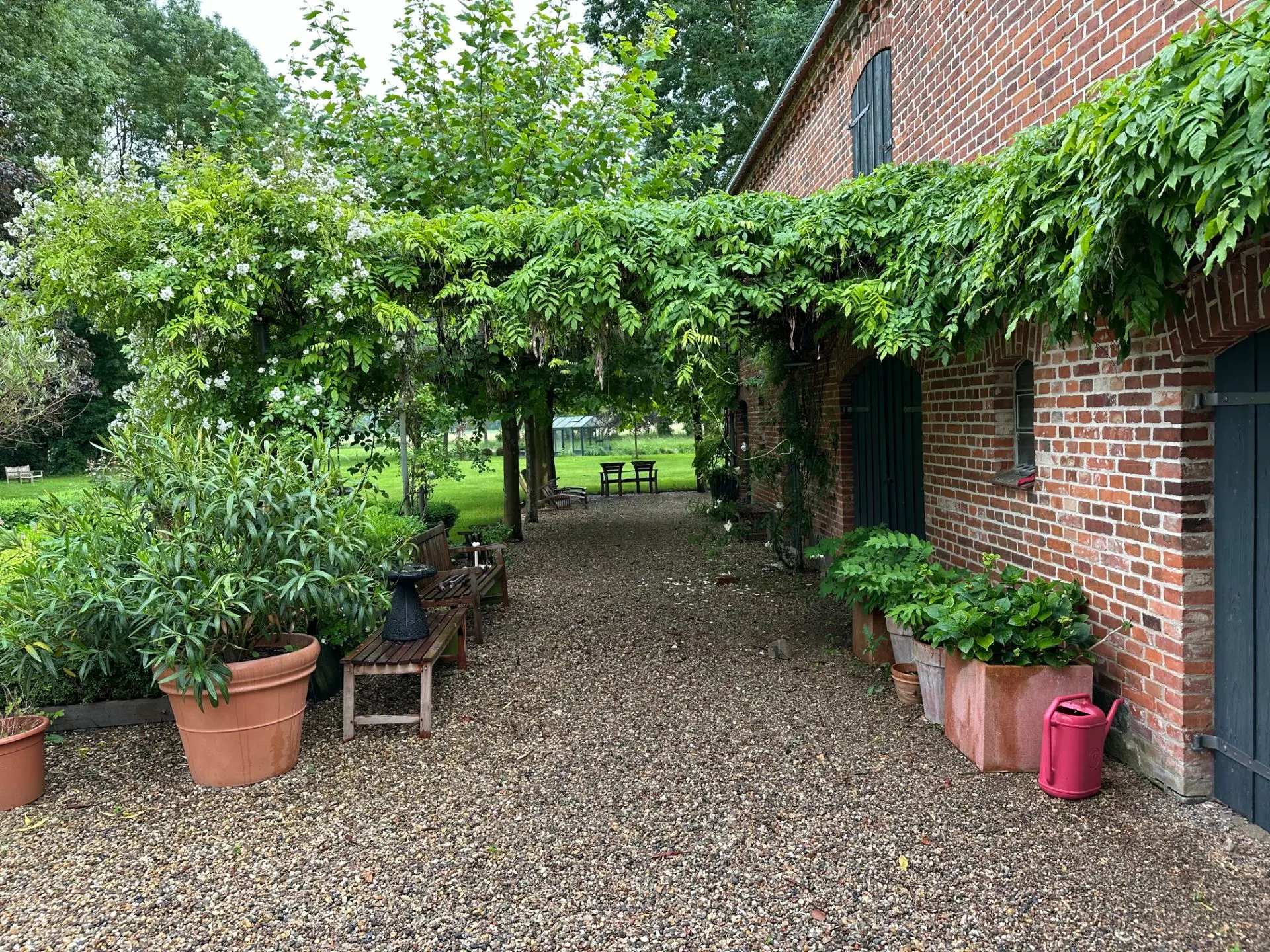 Gepflasterter Weg neben Backsteinhaus mit Pflanzen in Töpfen und grüner Überdachung.