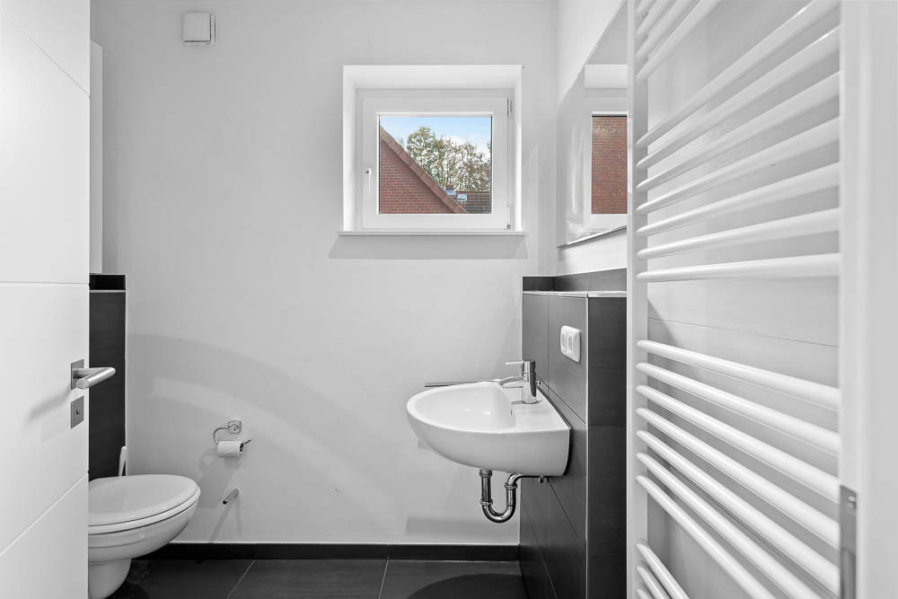 Helles Badezimmer mit Wandtoilette, Waschbecken, Fenster und weißem Heizkörper.