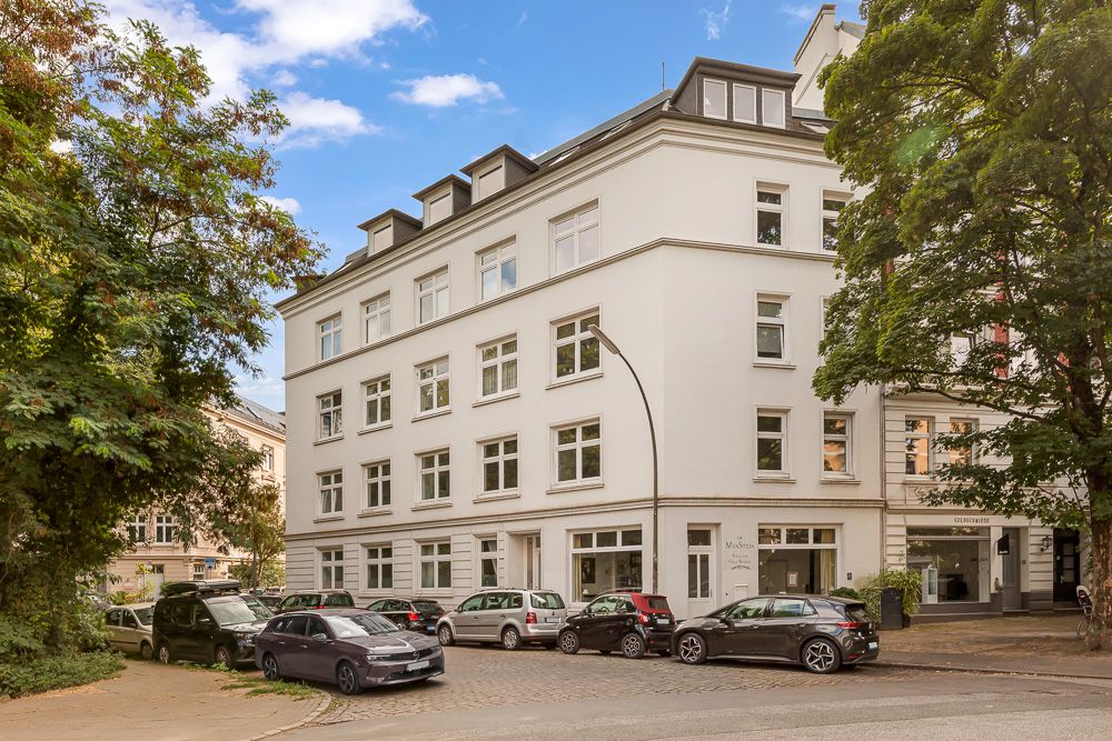 Mehrstöckiges weißes Wohngebäude an einer Straßenecke mit parkenden Autos und Bäumen.