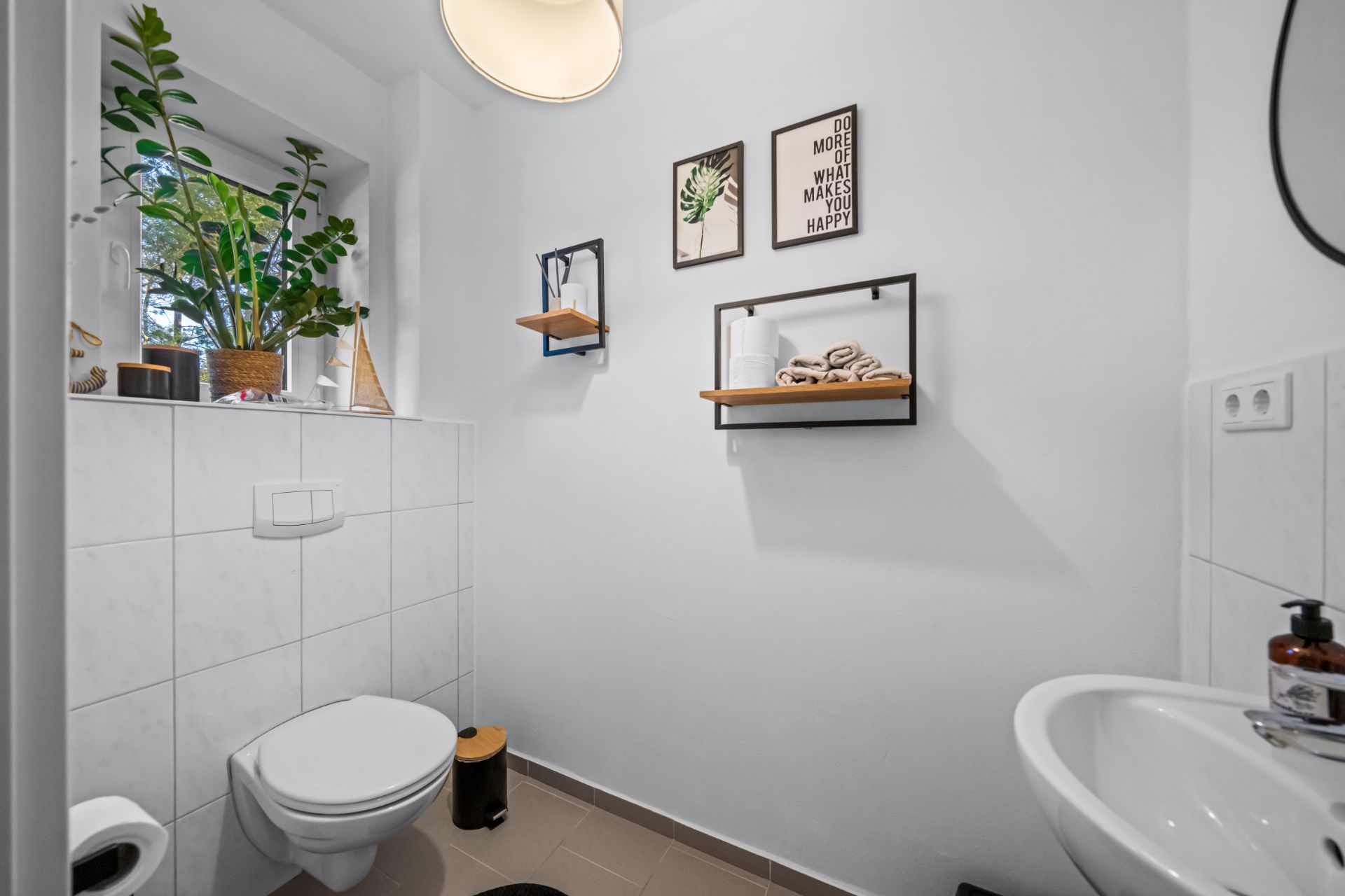 Modernes Badezimmer mit Wandtoilette, Waschbecken, Pflanzen und Regalen an weißer Wand.